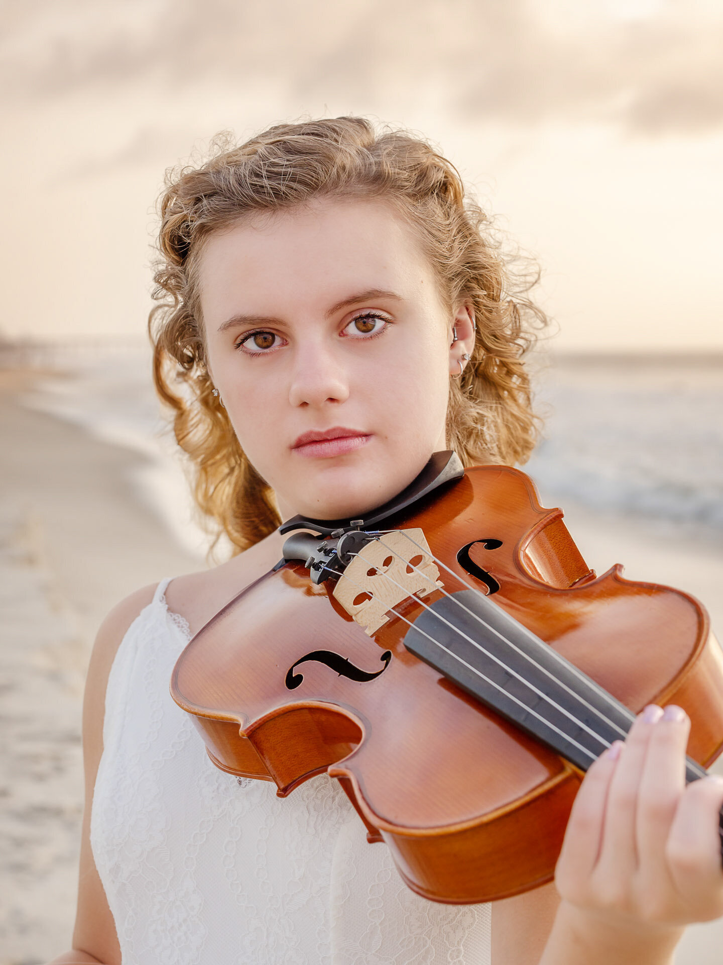 A high school senior in a white dress holding a viola, standing on the beach with the ocean as a backdrop in North Carolina. This close-up portrait highlights musical talent, perfect for unique senior photography in Wilmington, NC.