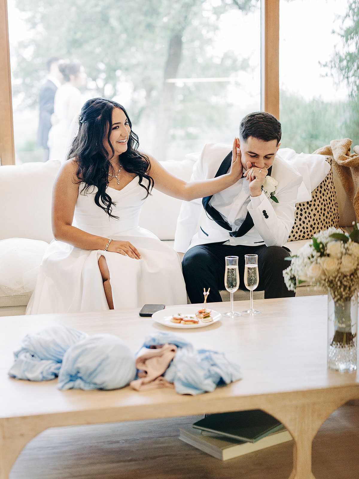 A bride in a white dress gently touches her smiling groom’s face as they sit on a couch. Captured by an Eau Claire Wedding Photographer, champagne glasses, a bouquet, and appetizers rest on the wooden table before them.