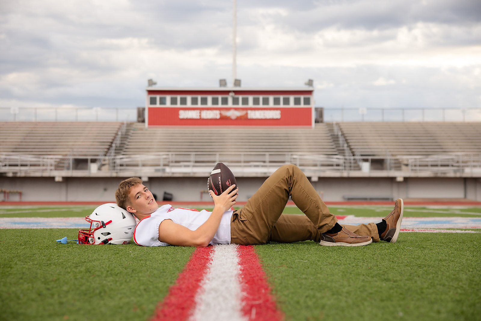 senior boy with football lying on turf with stadium in background