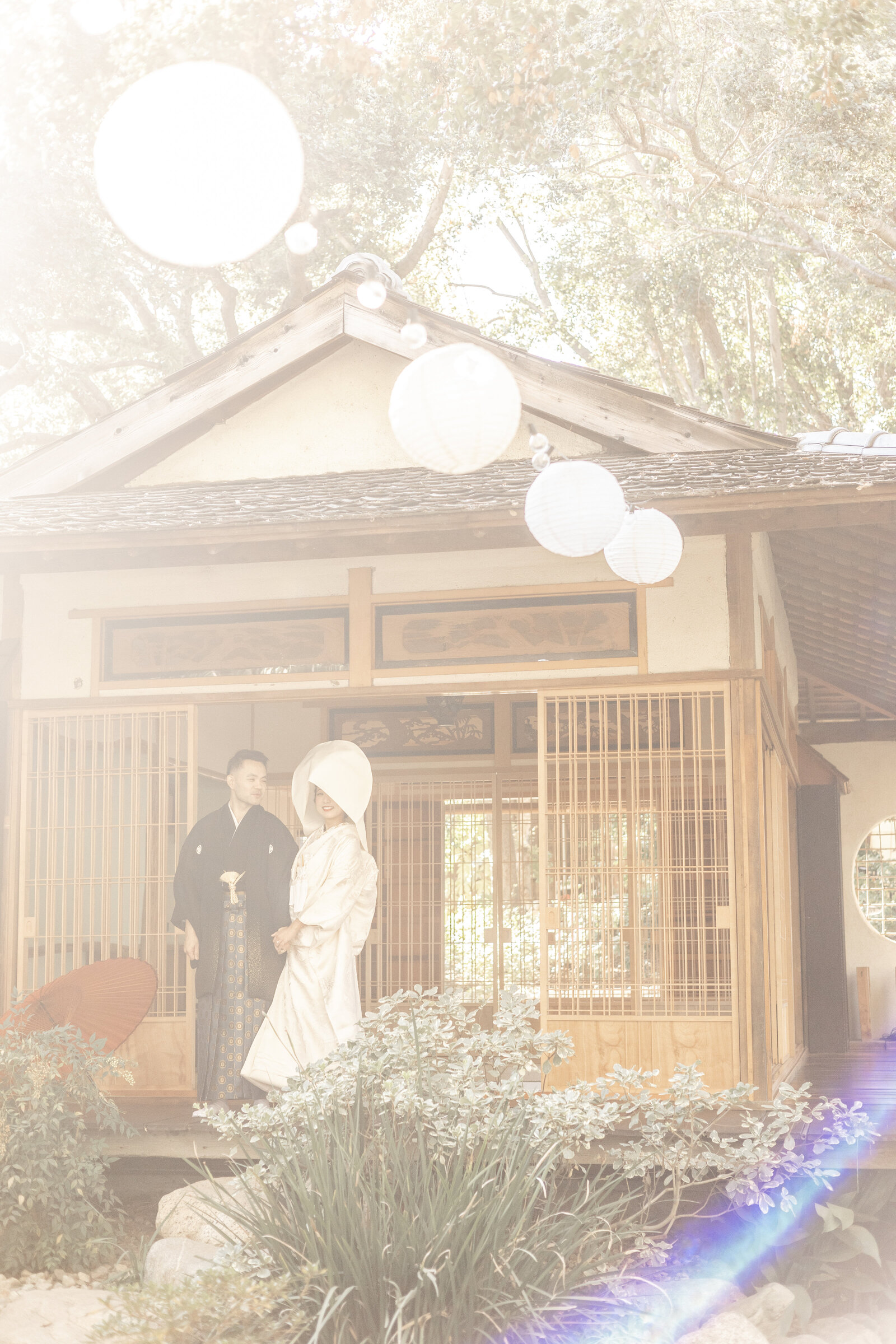 Bride and groom standing in front of the tea house at Storrier Stearns Japanese Garden in Los Angeles, with softly glowing paper lanterns above. Bride wears traditional white shiromuku and wataboshi. Available for kimono rental in Los Angeles | 提灯が柔らかく灯るロサンゼルスのストーリア・スターンズ日本庭園の茶室前に立つ白無垢と綿帽子の花嫁と新郎。着物レンタル可能。