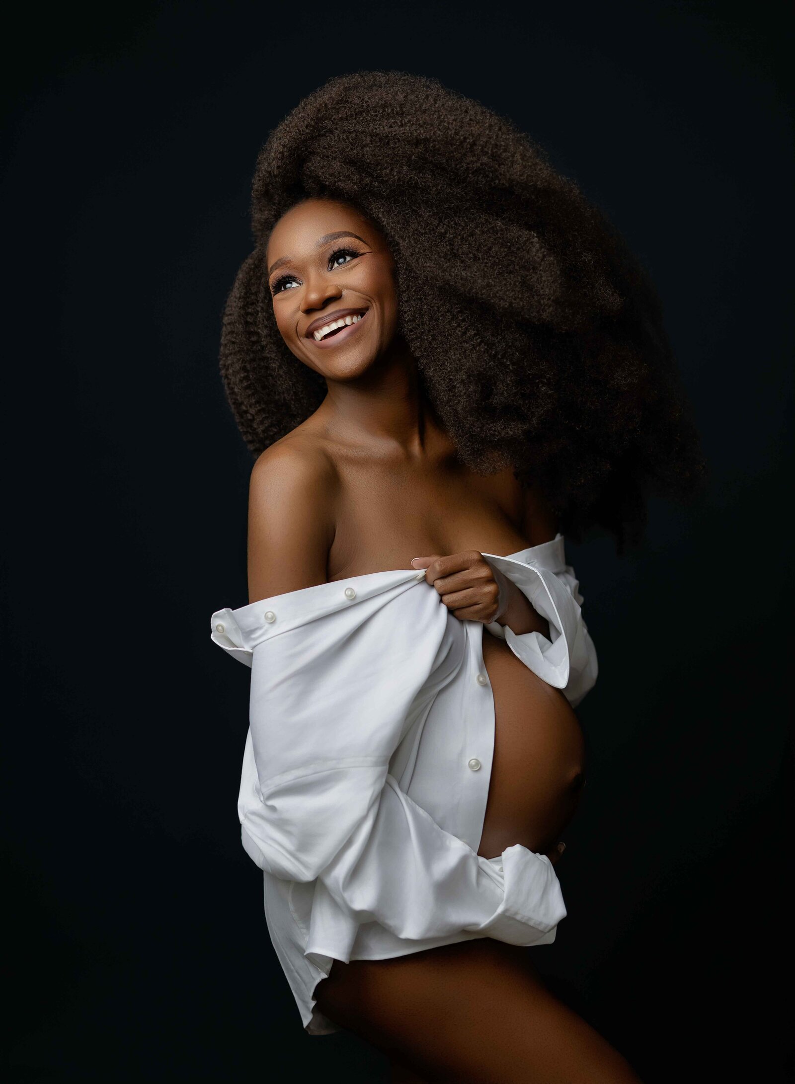 Pregnant women wearing a men's button don shirt and showing of baby bump while laughing over her shoulder. Photographed by Atlanta Maternity Photographer.