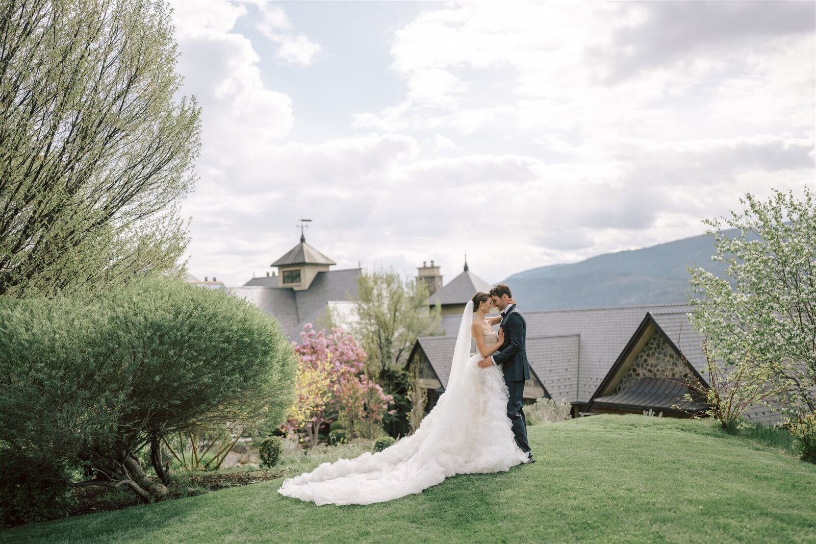 A wedding couple standing on the grass, hugging, with pink magnolia trees blooming and the Chateau Okanagan wedding venue in the background