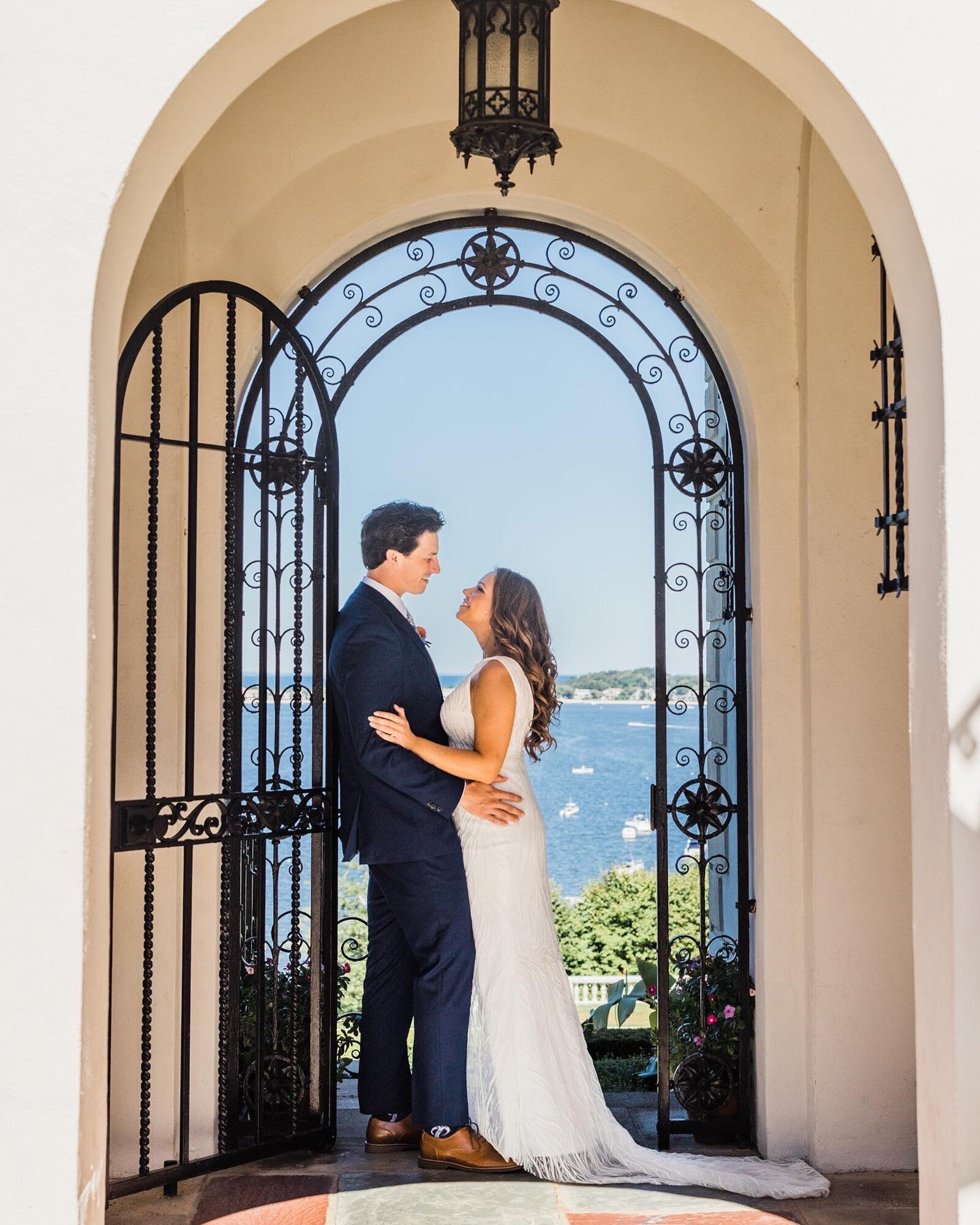 bride and groom stand under an ornate iron archway, facing each other and smiling