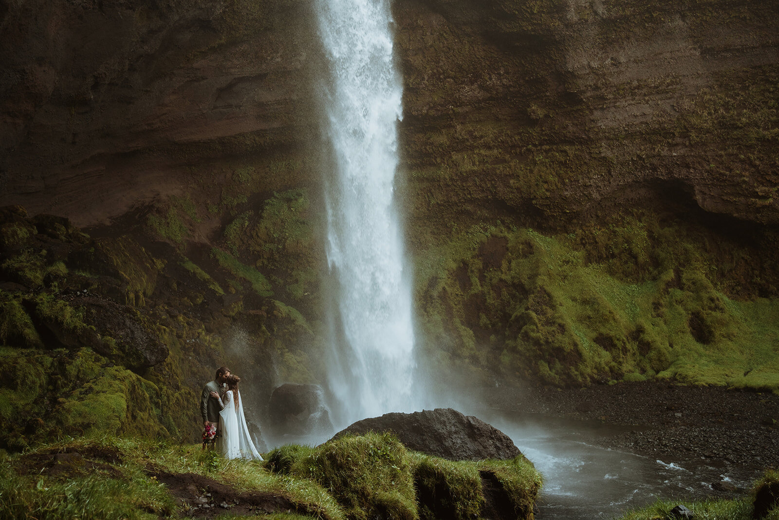 the bride and groom are at the base of a waterfall in greenery. they are kissing each other as the waterfall is cascading down behind them in the columbia river gorge.