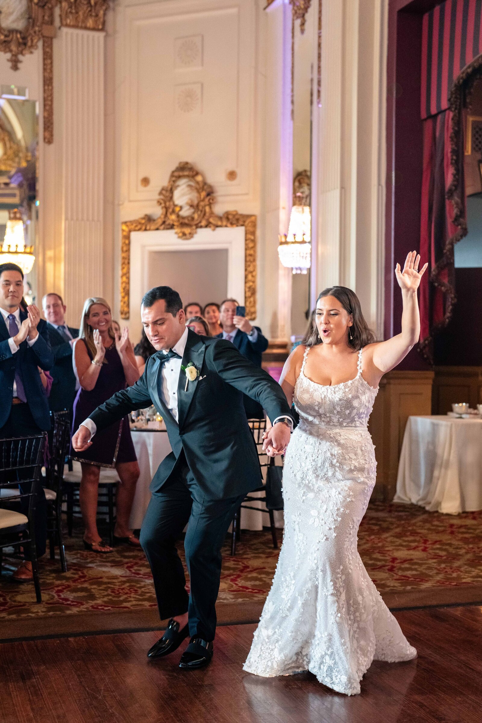 A bride and groom dance joyfully at their wedding reception. The bride wears a white lace gown, and the groom is in a black tuxedo. Guests are clapping and cheering in the elegant ballroom setting.