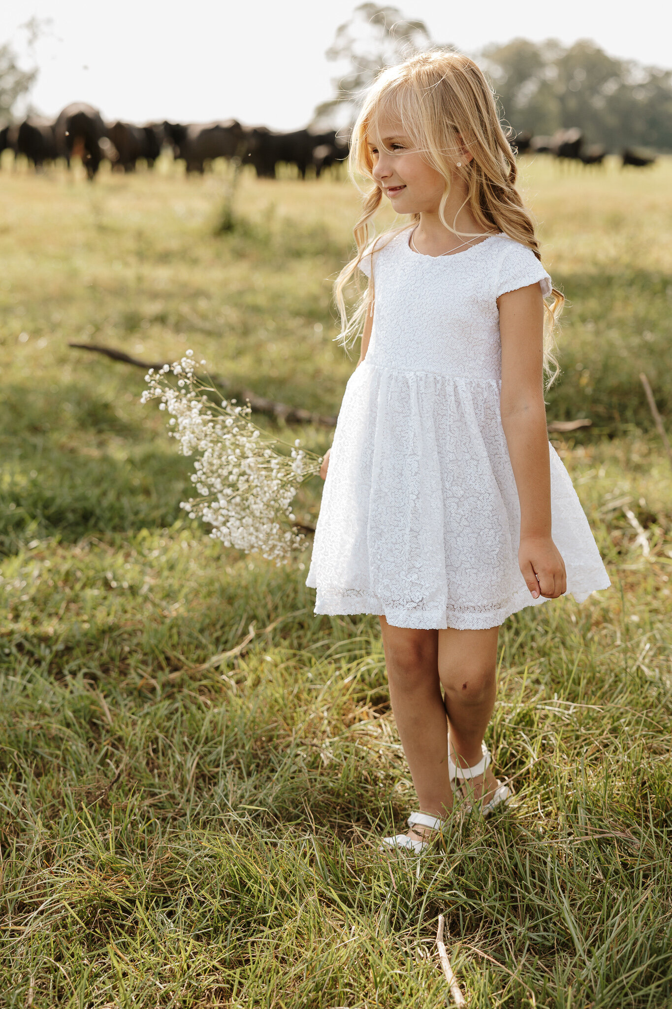 Little girl in a white dress walking through tall grass during a children's session in a farm field in Aiken SC.