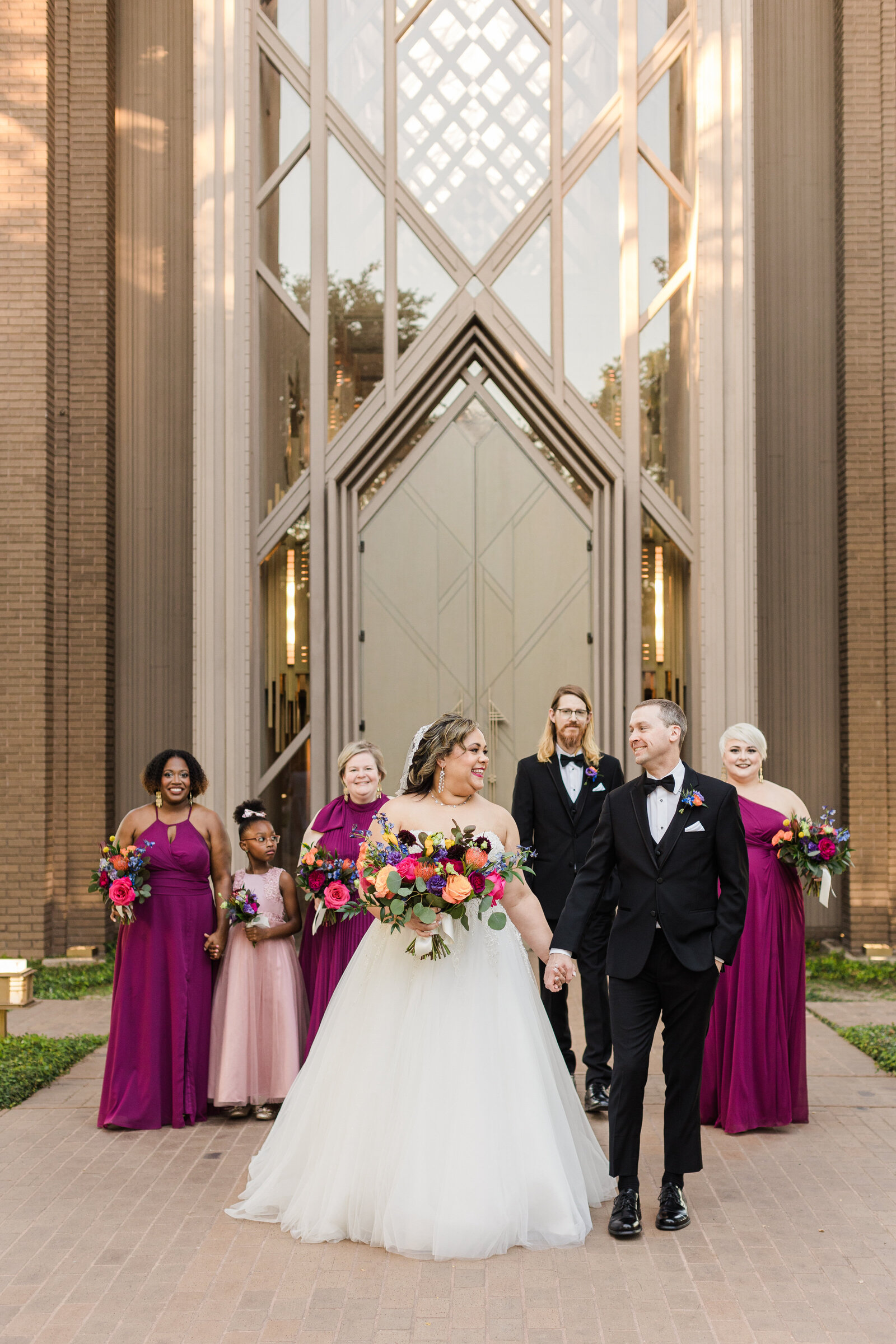 A bride and groom smiling and looking at each other alongside their wedding party in front of the Marty Leonard Chapel in Fort Worth, Texas. The bride is on the left and is wearing a long white dress and holding a bouquet. The groom is on the right and is wearing a black tuxedo with a boutonniere. Their wedding party is wearing either purple dresses or a black tuxedo.