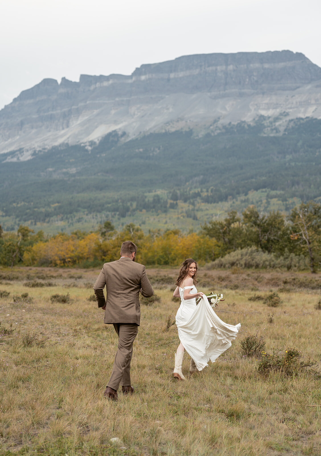 A bride looks back smiling while running through a golden Montana field with her groom, surrounded by mountain cliffs and fall foliage, captured by Sydney Breann Photography during their Glacier National Park elopement.
