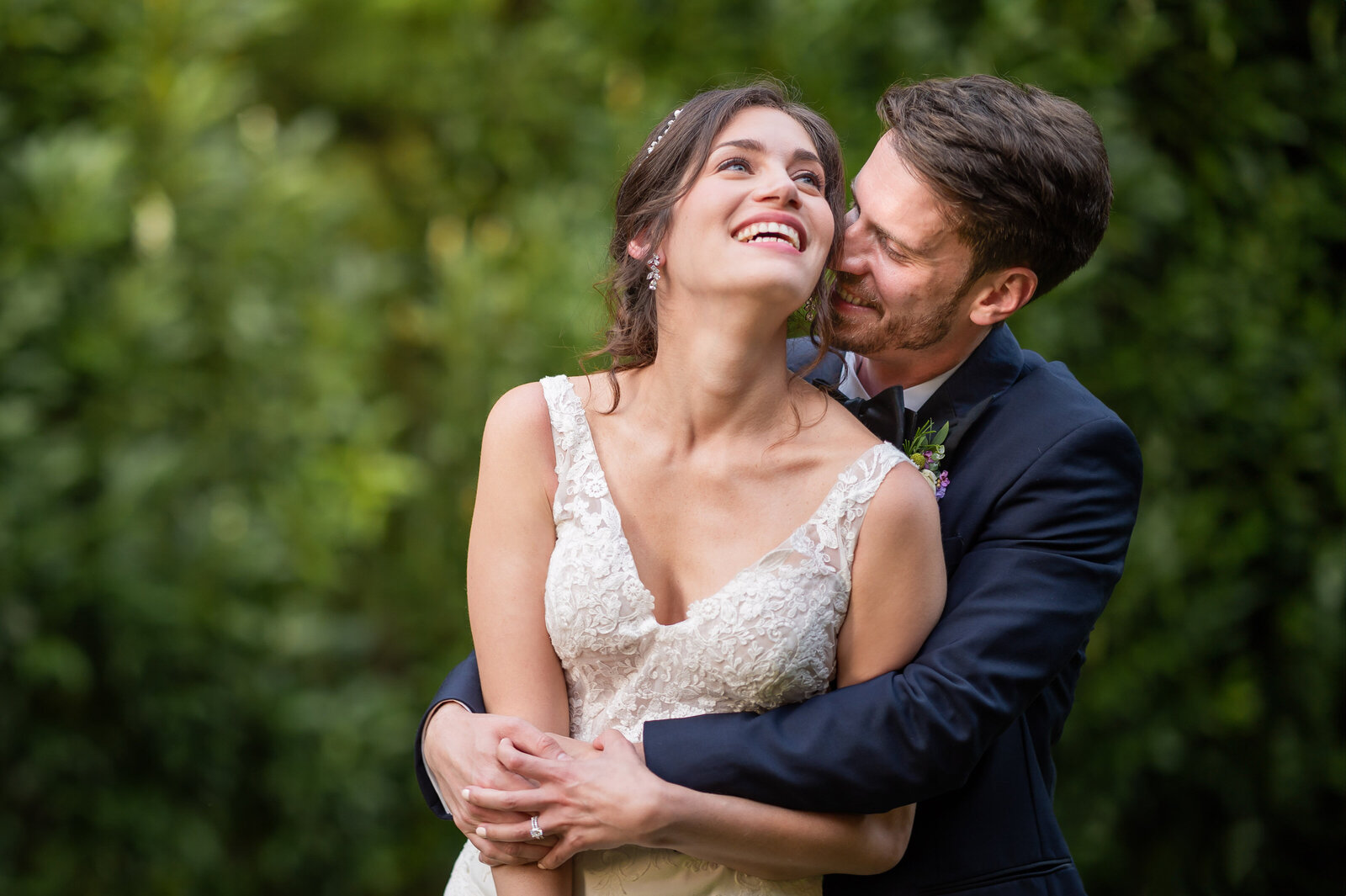 A couple in formal attire embraces outdoors, with the man behind the woman. She smiles while he leans forward, surrounded by a green, leafy backdrop.
