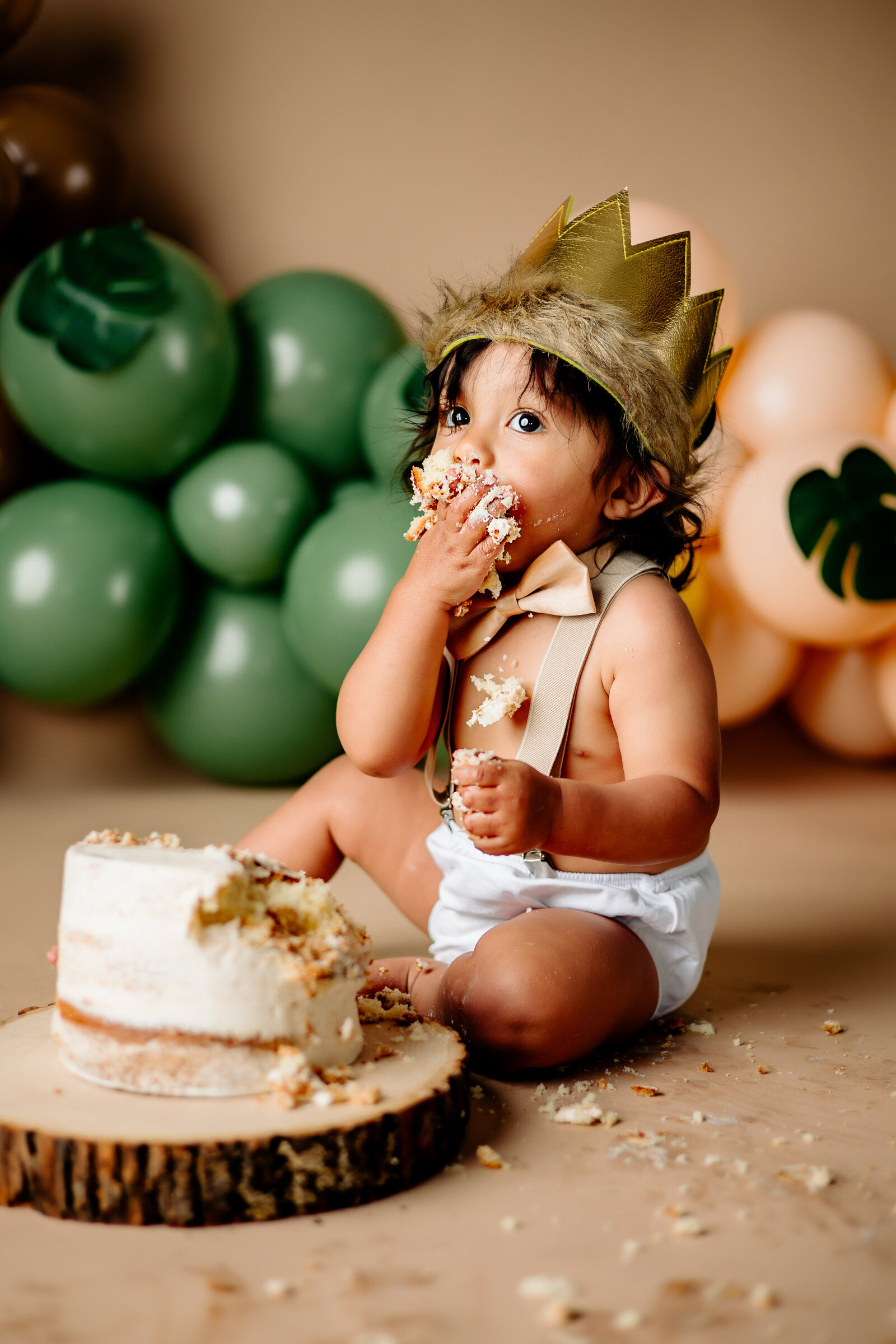 The messy aftermath of a cake smash , with crumbs and frosting  scattered around a happy baby smashing one last bite into his mouth