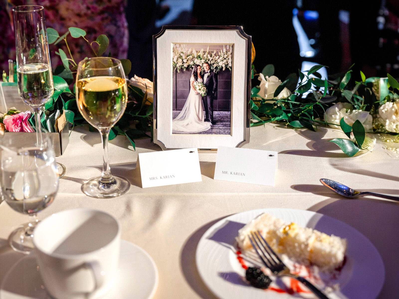 A wedding reception table shows a framed photo of a couple in wedding attire. Nearby, there are two glasses of wine, floral decorations, place cards, and a plate with a slice of cake.