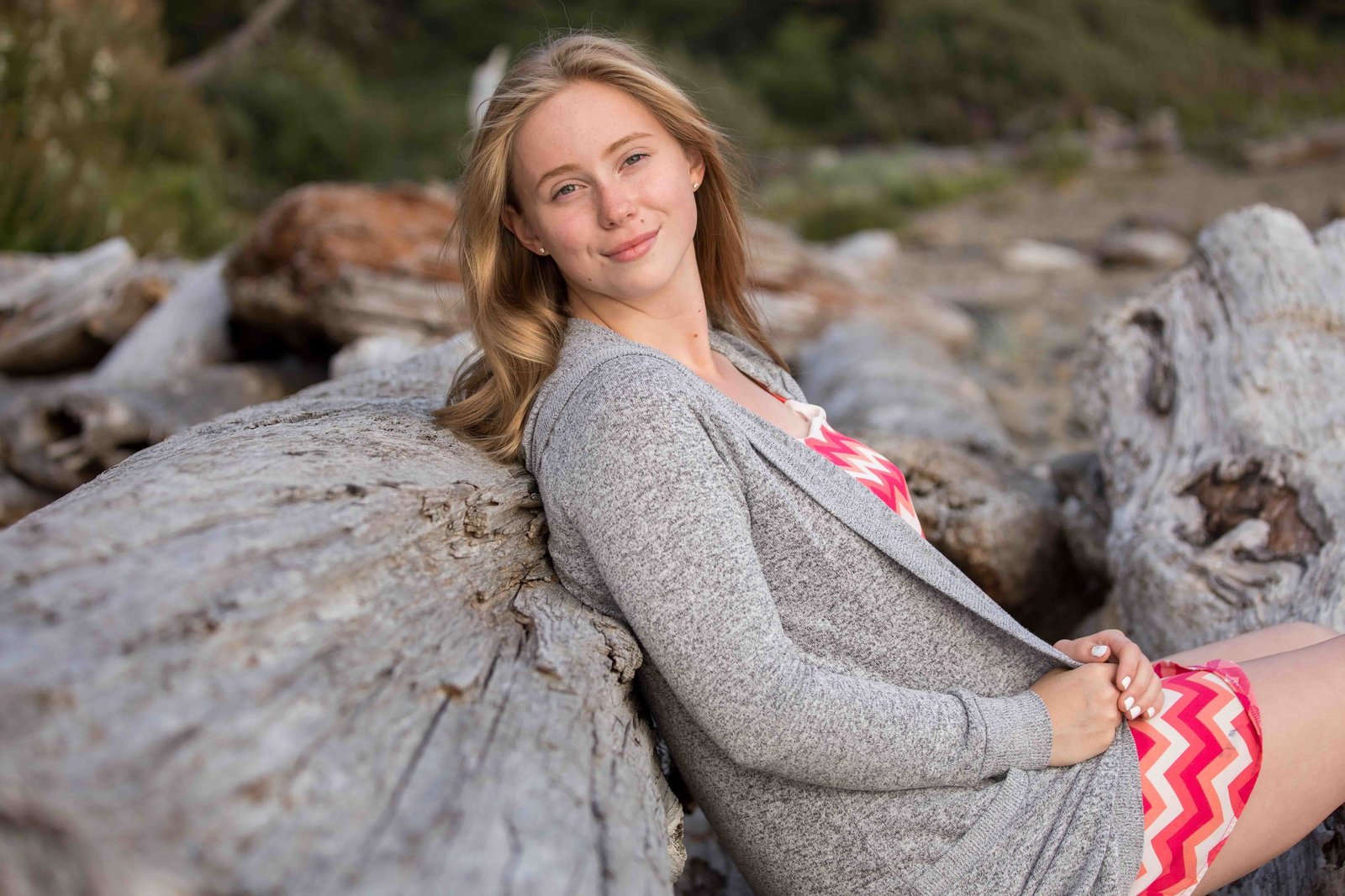 A young girl has her back to a piece of driftwood. She has a small smile on her face this was her senior photo session with senior photographer JodyRae. They are at hug point State Park and she is wearing a pink dress with white stripes