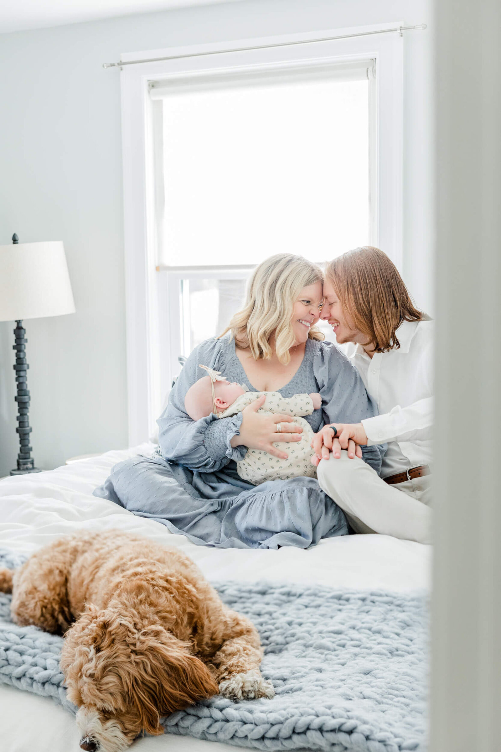 Mom, holding a sleeping newborn, and dad smile and put their foreheads together while a dog lays on a knitted blanket on a bed