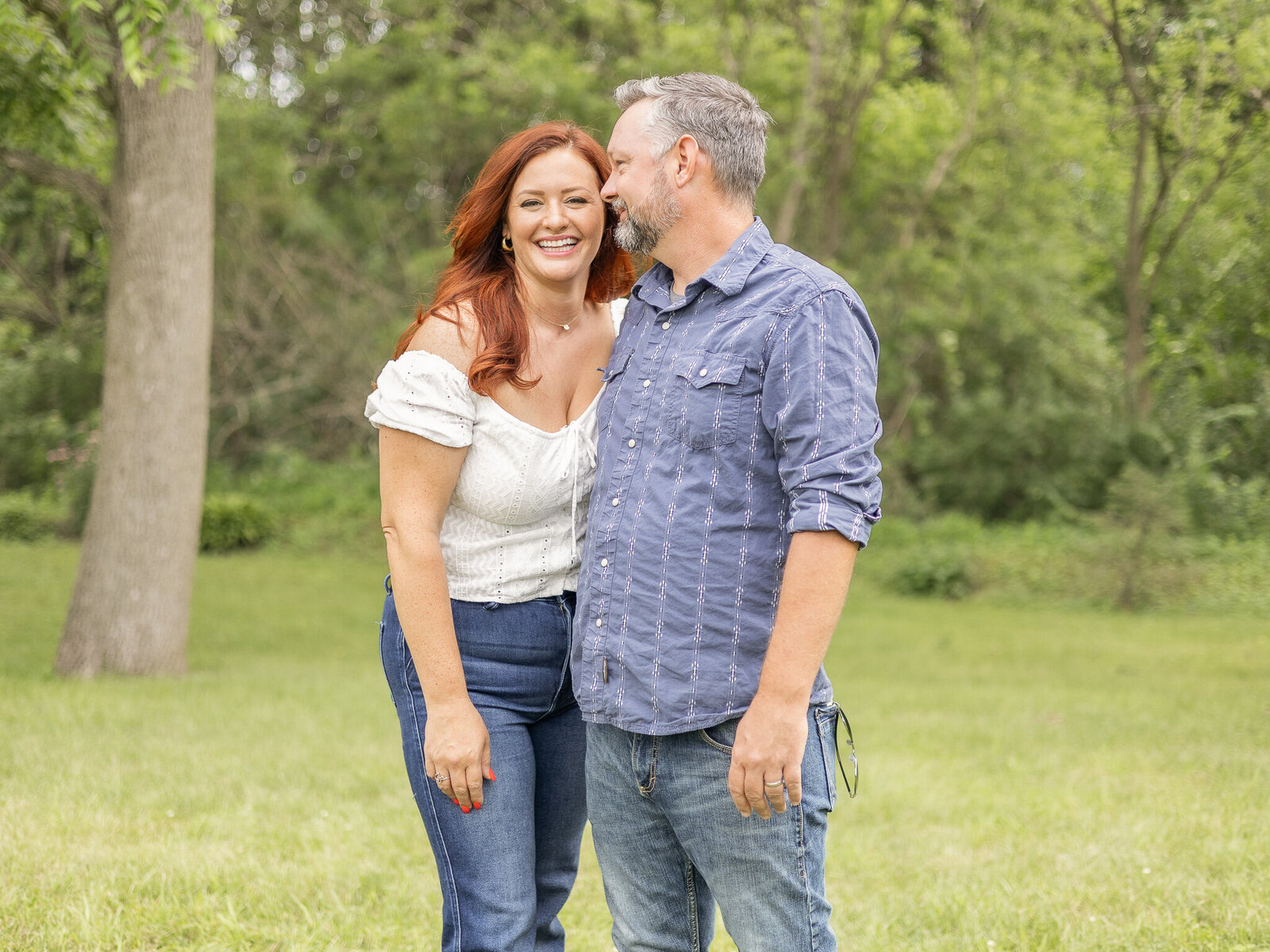 Husband and wife standing in grass with a tree line behind them.  Wife has red hair, wearing a white shirt and blue jeans, looking at the camera and laughing.  Husband is next to her whispering in her ear, with pepper hair wearing a white and blue shirt and blue jeans.