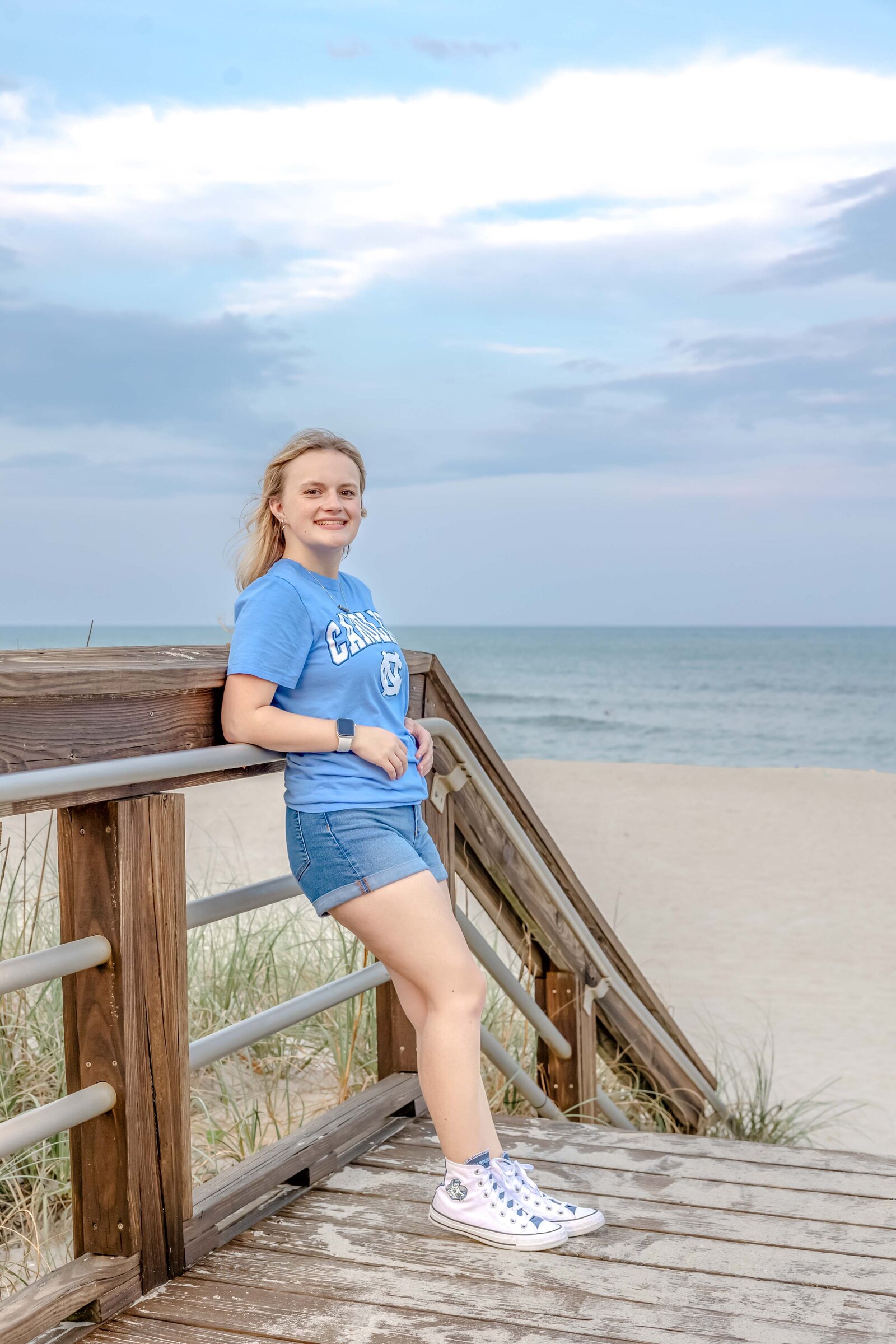  A high school senior in a carolina blue UNC shirt and shorts leaning on a boardwalk railing with the ocean in the background in Carolina Beach, North Carolina. This casual and vibrant portrait captures the relaxed vibe of senior photography, perfect for beach-themed sessions.