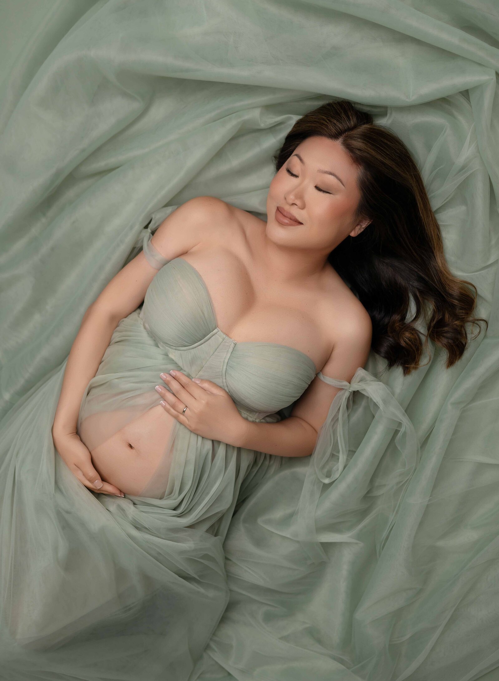 Pregnant women with dark hair, lying on tulle gown and holding baby bump.