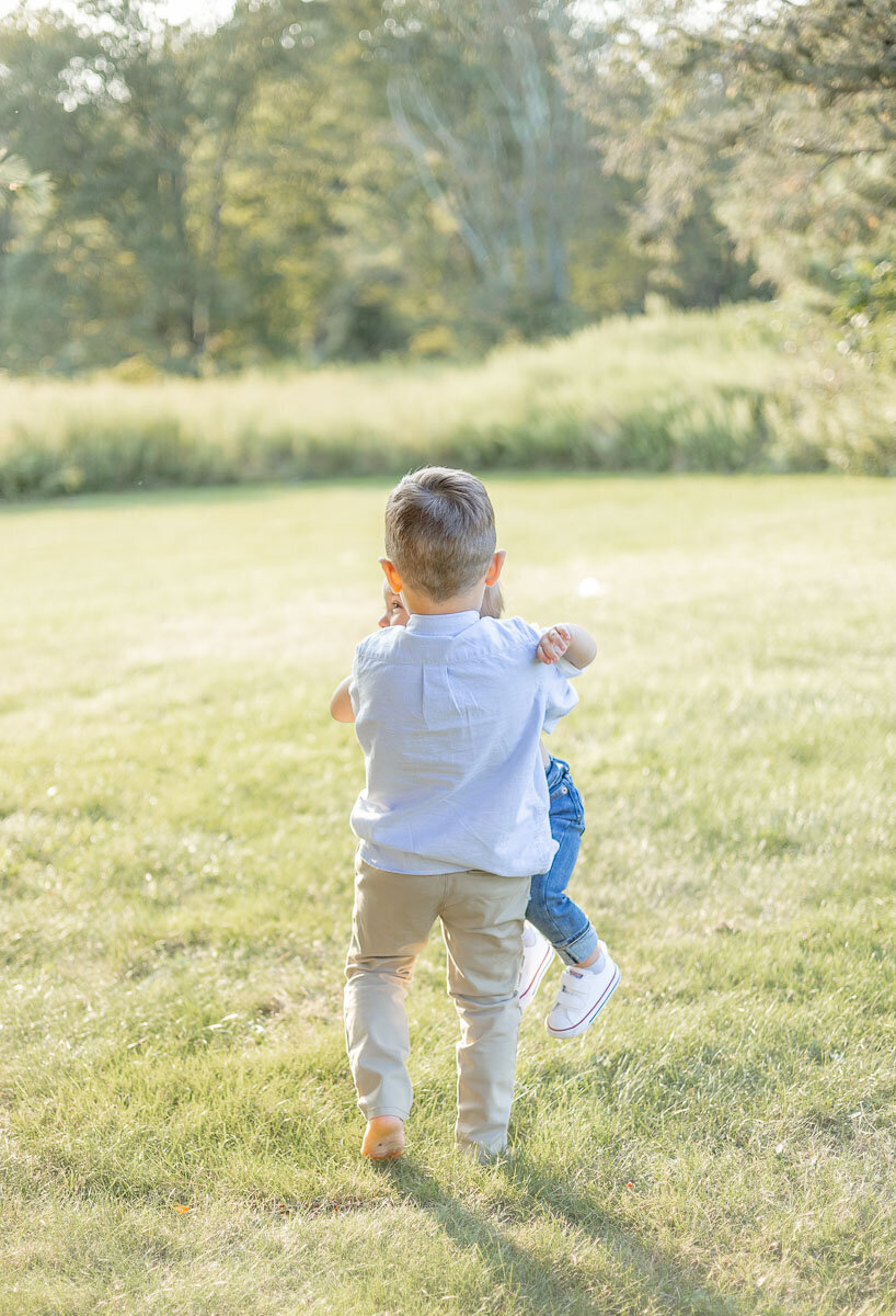 Older brother holding his toddler sibling during golden hour in a sun-drenched field—captured by Nicole, a lifestyle family photographer serving Westchester and Connecticut families.