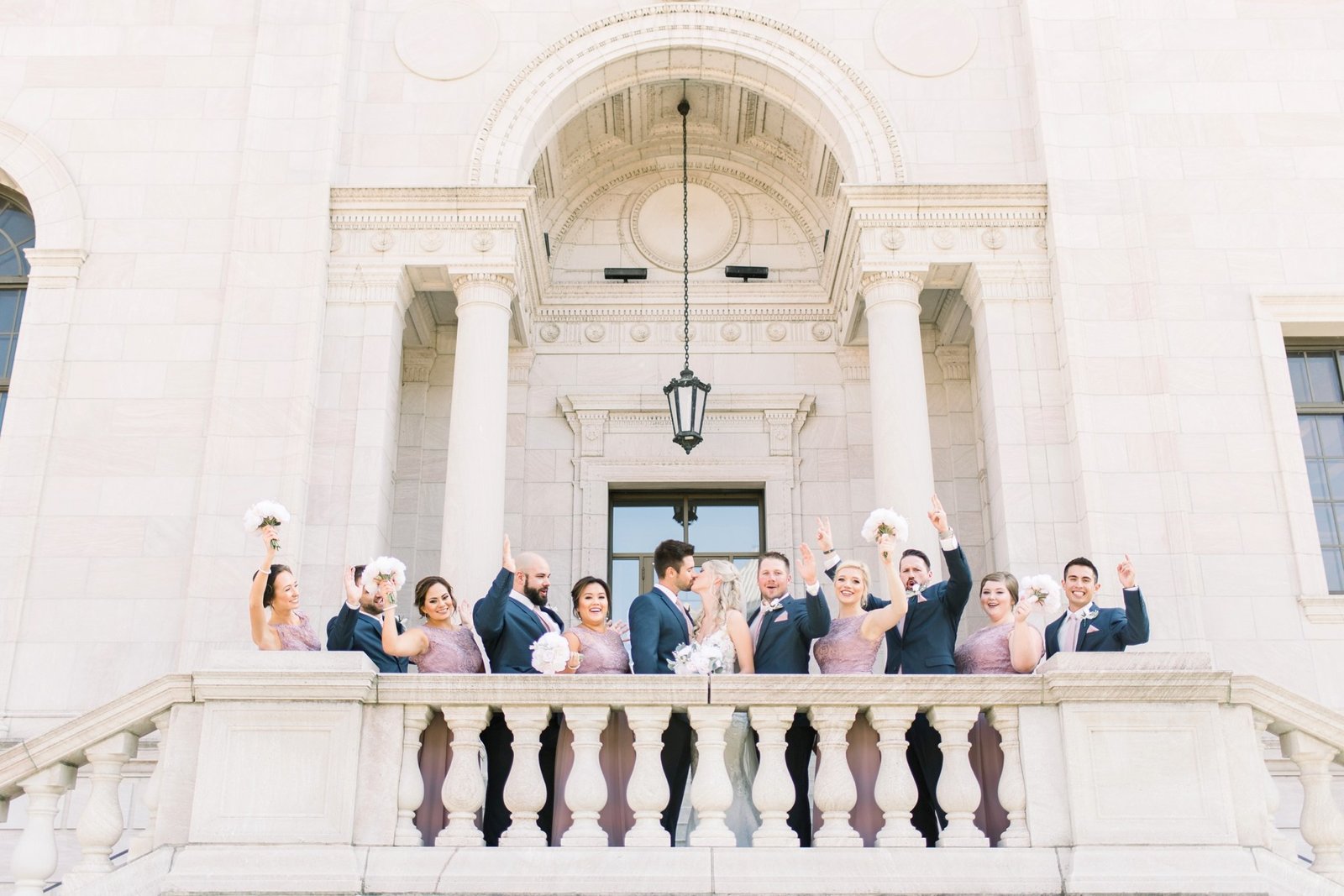 A wedding party (wearing purple and navy) cheer on the bride and groom as their kiss in the balcony of the James J. Hill House before their Landmark Center wedding in St. Paul, Minnesota.