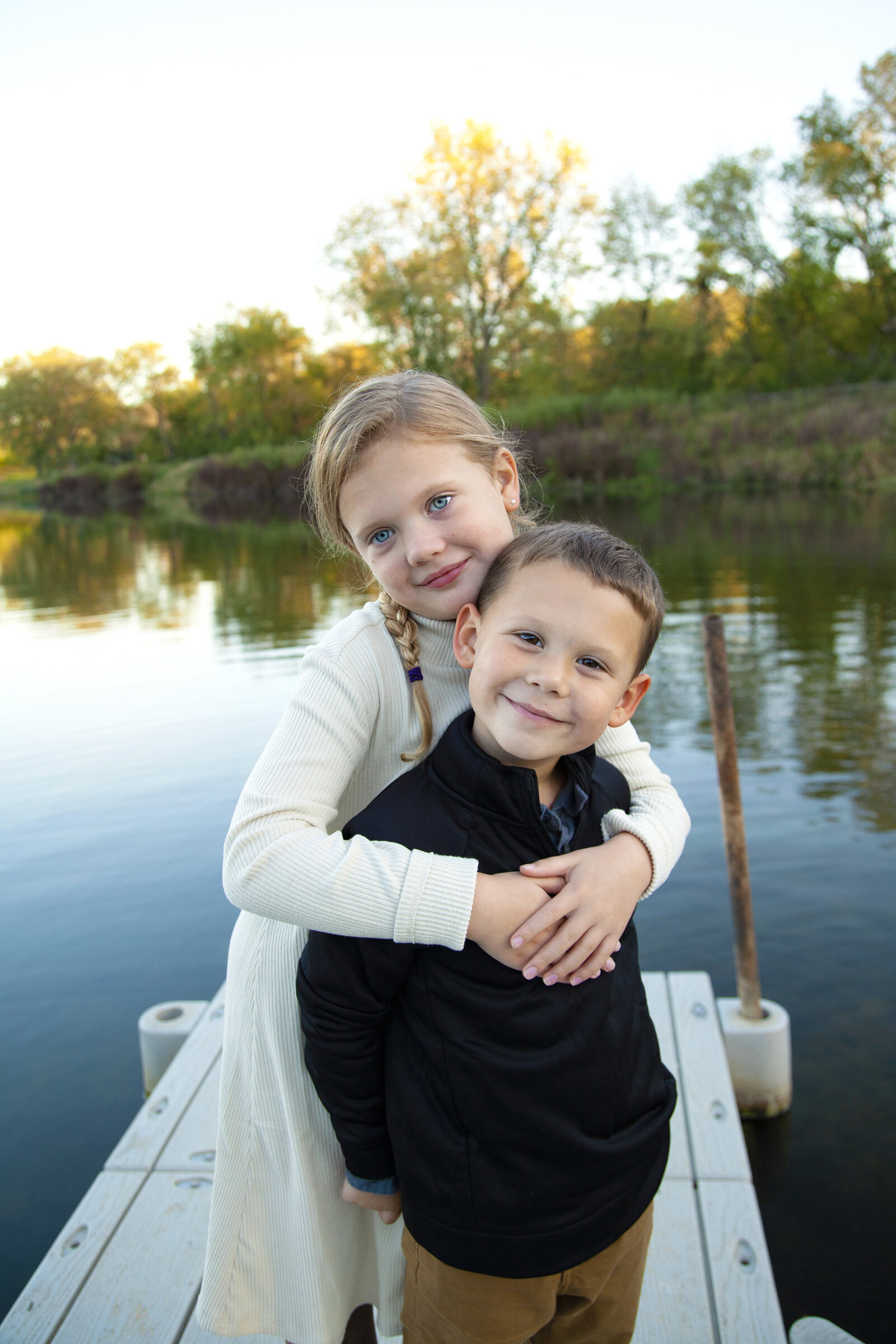Sister hugging brother around shoulders on dock in front of pond.