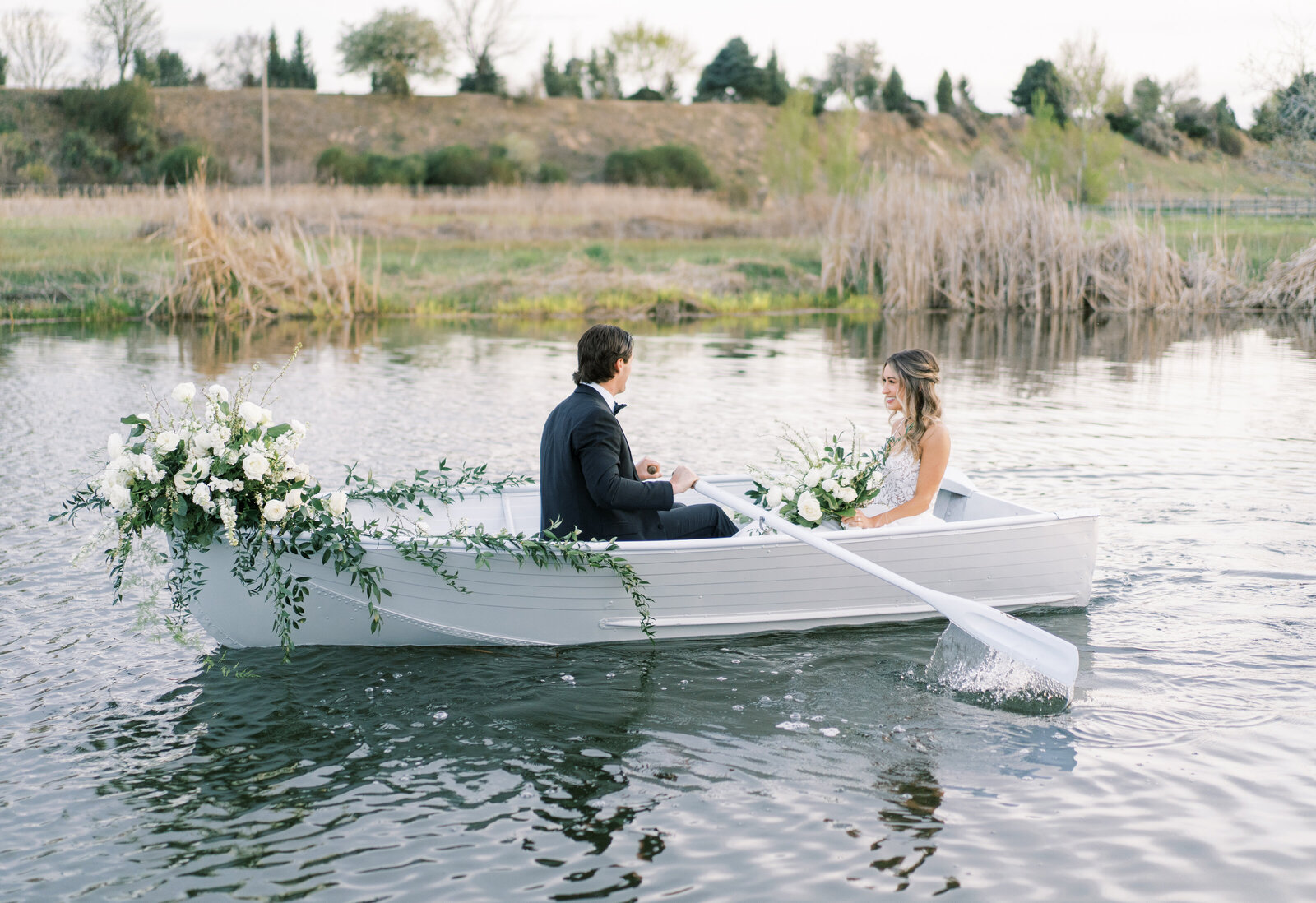Portrait of a bride and groom in a white wedding gown and black tuxedo in a boat filled with flowers and greenery on a lake.