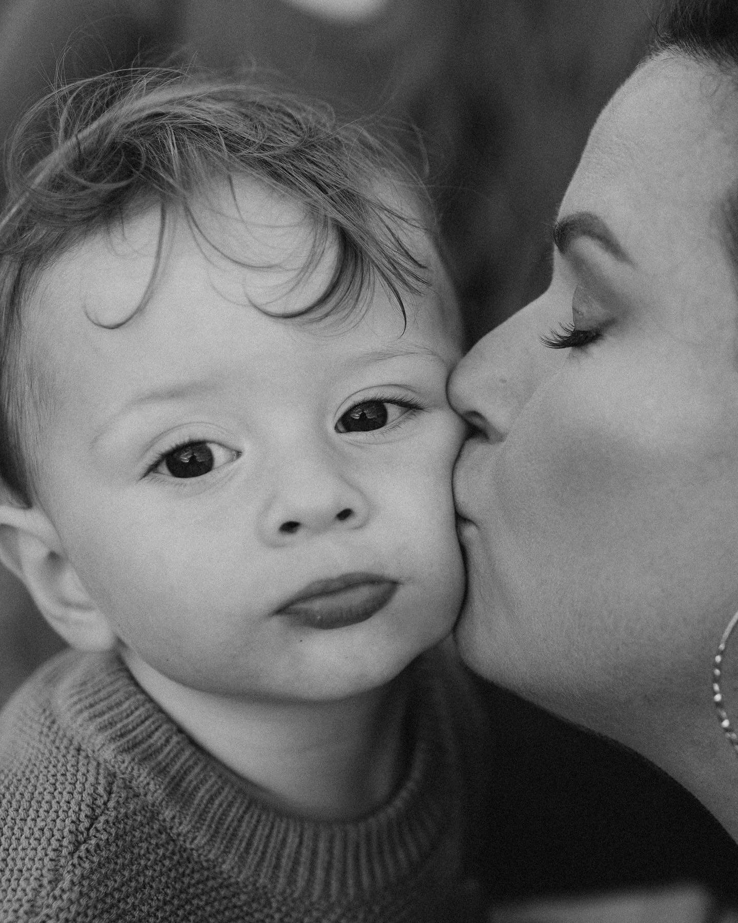 A black and white photo of a woman kissing a young child on the cheek. the child, wearing a sweater, looks at the camera with a calm expression.