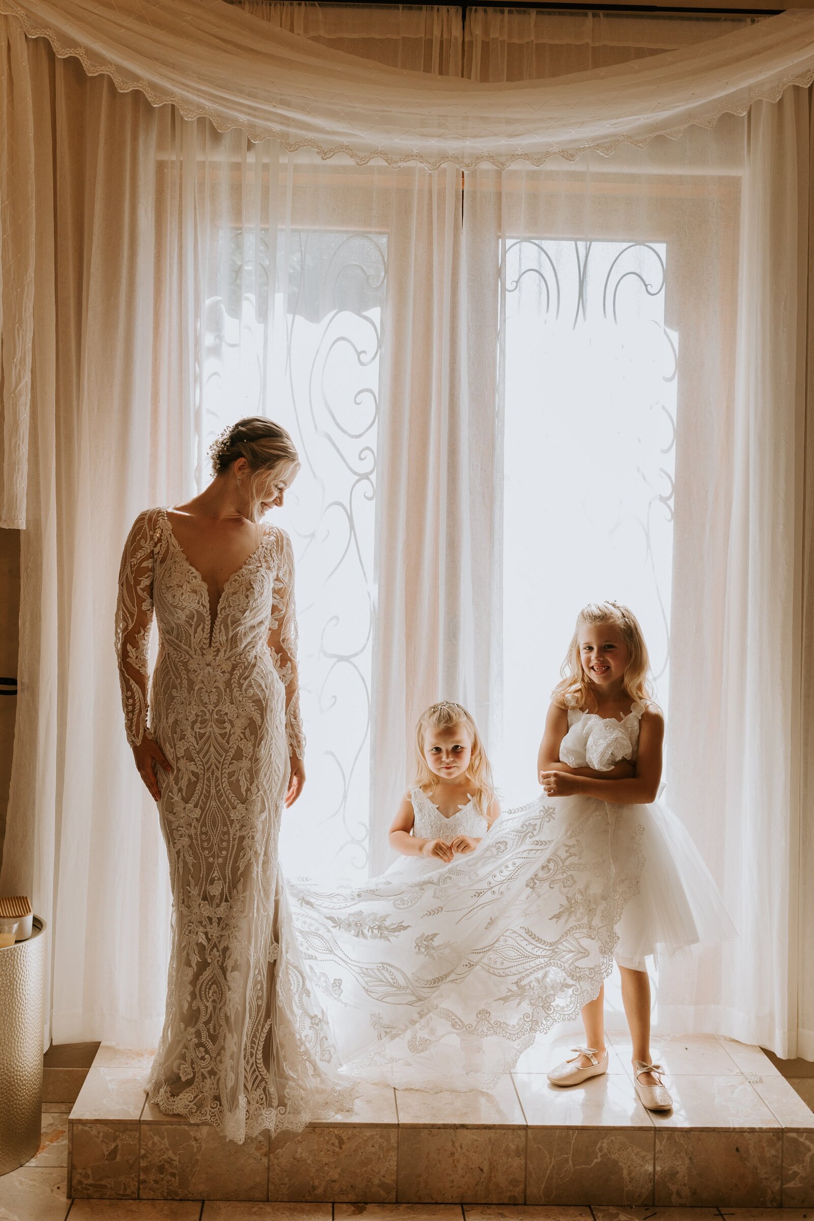 Bride stand in a beautifully lit room with her flower girls.
