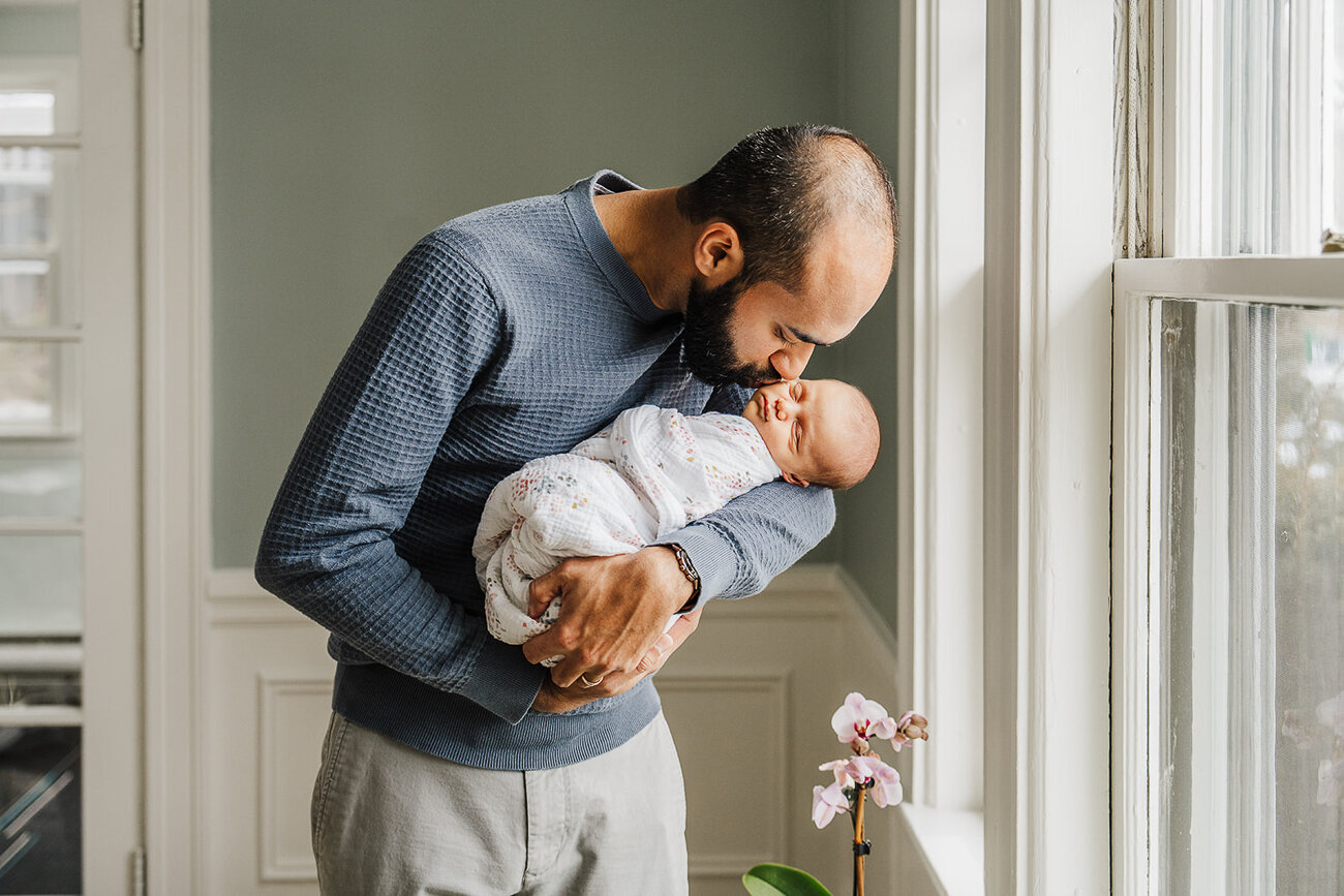 dad leans over new baby by window and kisses cheek