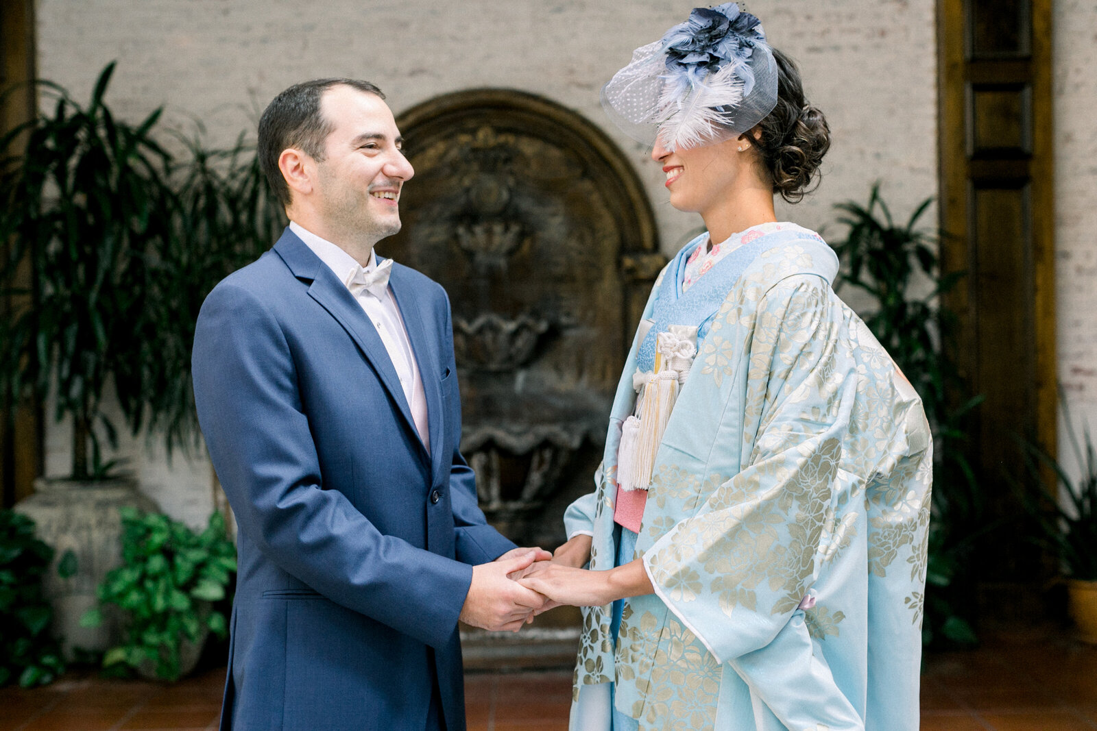 Bride in blue wedding kimono holding hands with groom in navy suit near a romantic fountain at The Ebell Club Long Beach, perfect for kimono rental in Los Angeles | ロサンゼルスのThe Ebell Club Long Beach噴水前でネイビースーツの新郎と手を取り合うブルーの着物レンタル花嫁