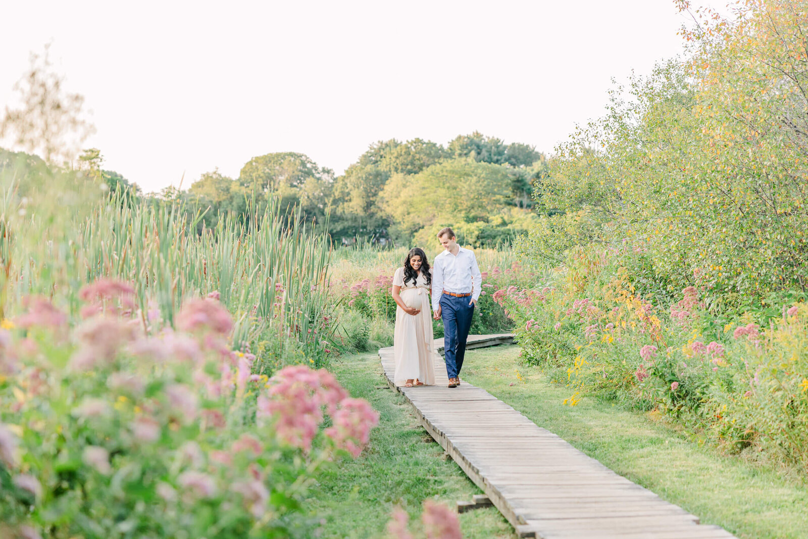 Pulled back view of a pregnant woman and her husband walking together on a wooden path between summer wildflowers, cattails and trees