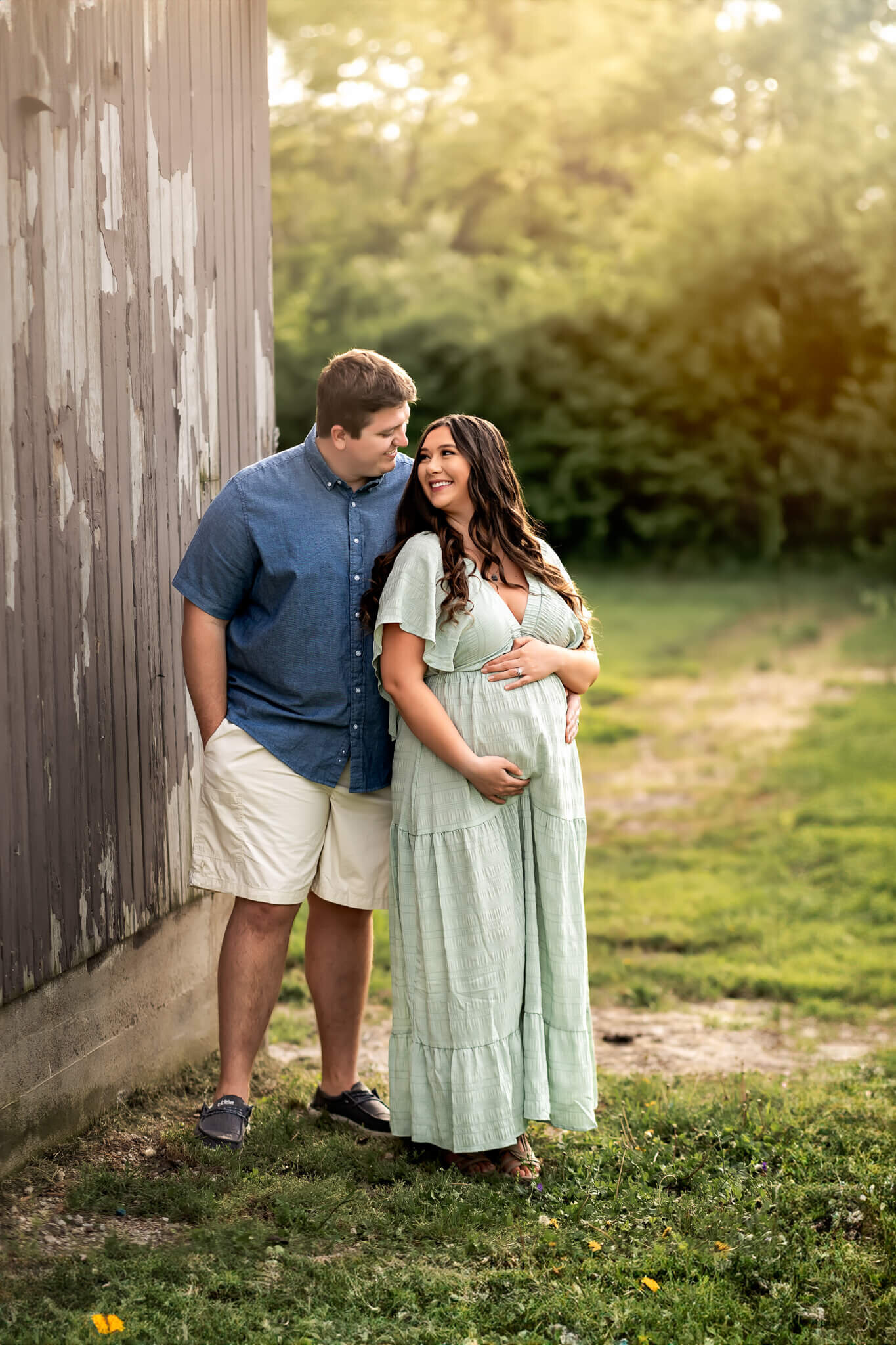A couple stands outside by a weathered wooden wall, smiling at each other. The woman in a light green dress cradles her pregnant belly as the man stands beside her. This sweet moment, captured by a Bloomington maternity photographer, glows with greenery and sunlight.