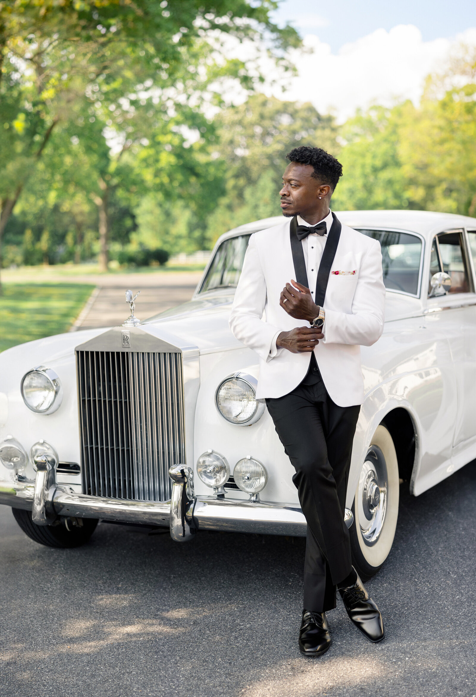 A stylish black man in a black tuxedo with a white jacket stands confidently in front of a classic white Rolls-Royce on a sunny day. Green trees and a clear blue sky are in the background. He is adjusting his watch and looking to the side.