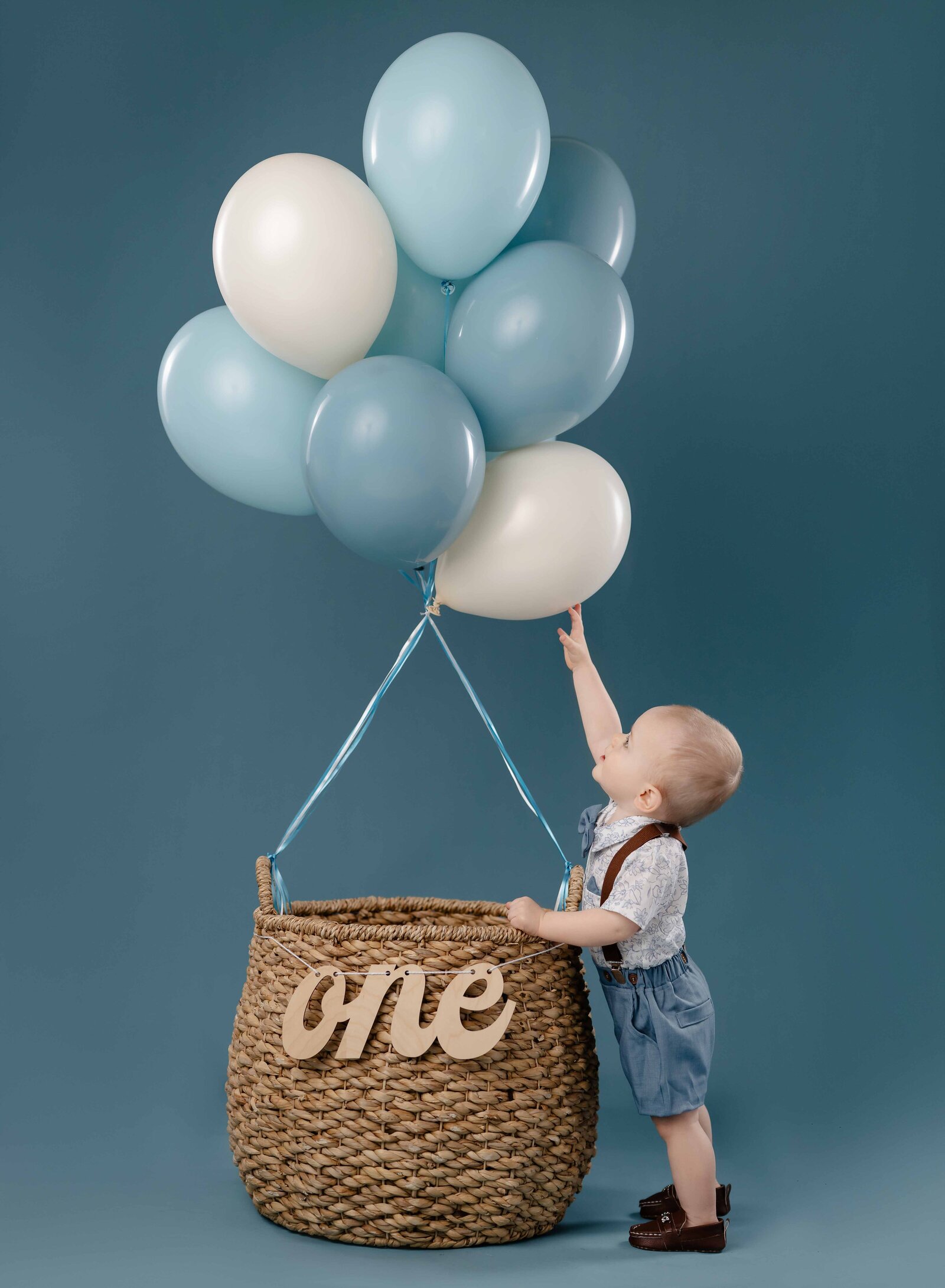 Baby standing next to balloon basket and reaching up to touch the balloons.