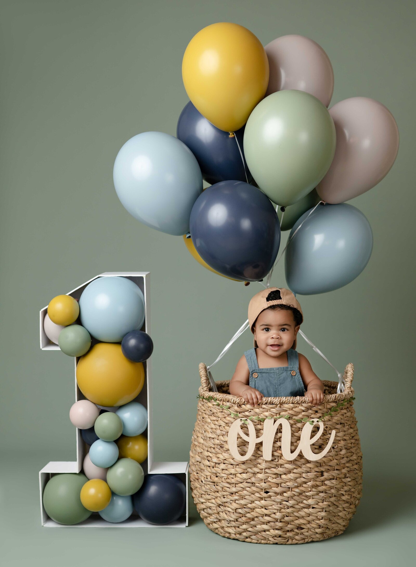 Birthday photo of baby boy standing in hot air balloon basket. Cake smash photo taken by Jenni Nikki Photography in Atlanta, GA.