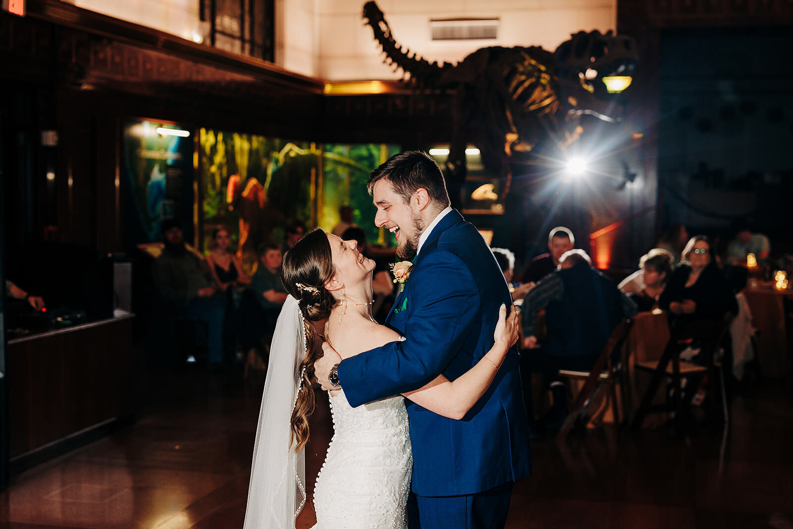 A bride and groom share their first dance in a museum in Downtown Austin, smiling at each other as a dinosaur skeleton looms in the background and guests watch from their tables.
