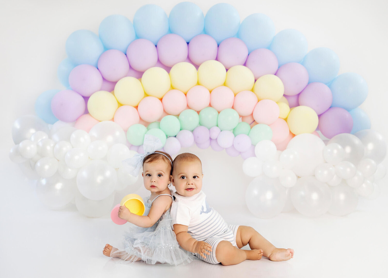 Two adorable babies sit against a white background with a colorful rainbow balloon arch behind them. One baby, wearing a light blue dress and headband, holds a yellow and pink toy while the other baby, dressed in a white outfit with blue accents, leans back on their hands. The balloon arch features pastel colors of blue, purple, yellow, pink, and green, creating a cheerful and whimsical scene.