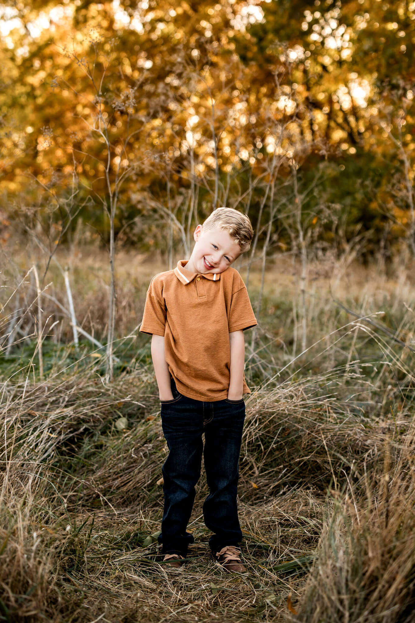Young boy with curly hair smiling, wearing a brown shirt and dark jeans, standing in a field with dry grass and autumn trees in the background. Captured as an outdoor portrait by a Bloomington family photographer, his hands are in pockets with head tilted slightly.