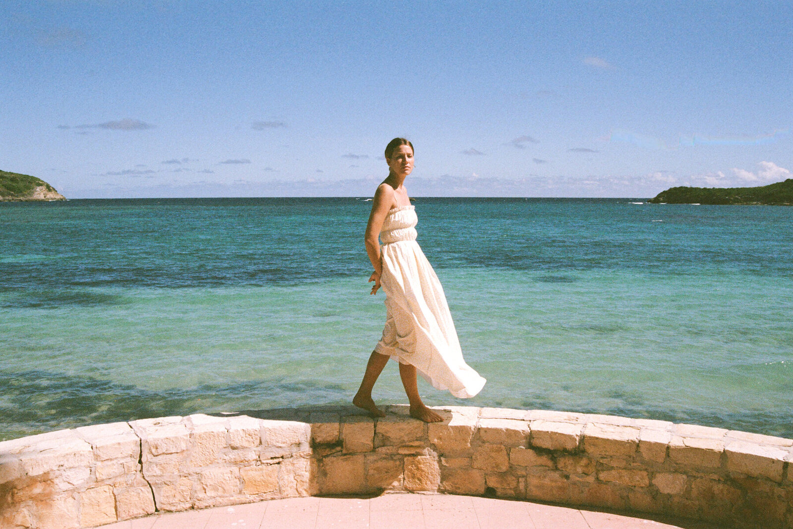 Female wearing white gauze strappy dress on stone wall with beach in background