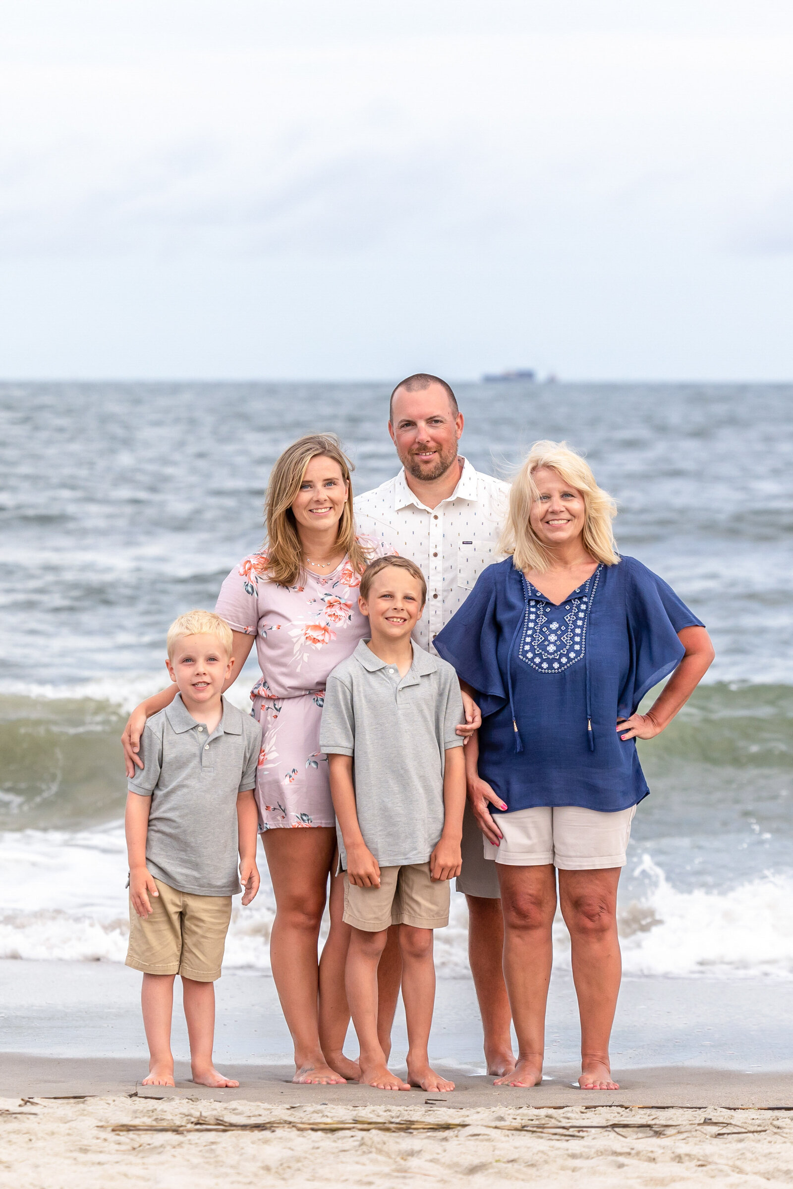 A family of five stands barefoot on Carolina Beach, dressed in casual summer attire. The ocean waves and overcast sky provide a classic beach atmosphere. Perfect for professional family photography in NC, highlighting close family connections.