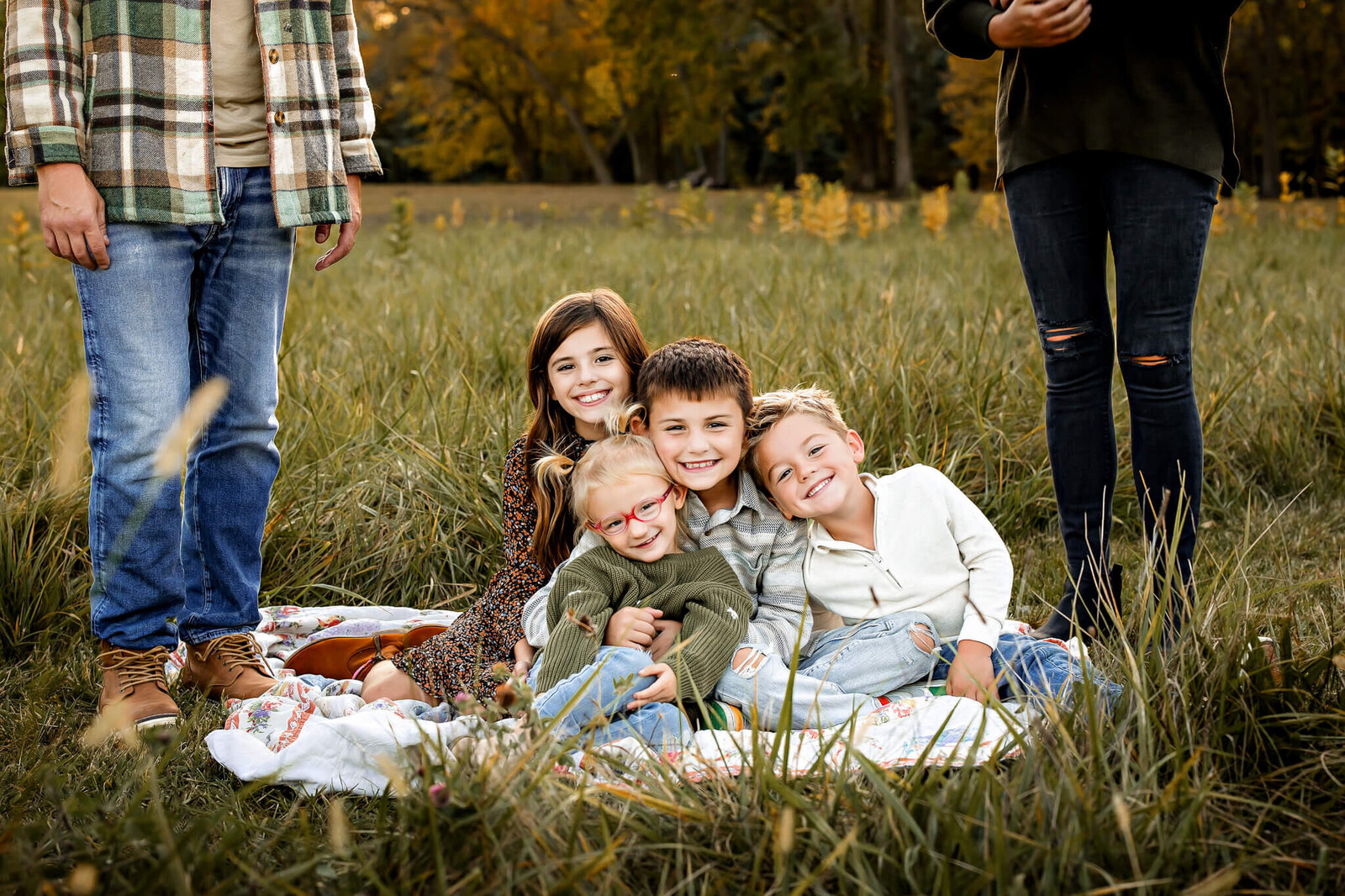 Four children are sitting on a blanket in a grassy field, smiling at the camera. Captured by a Bloomington family photographer, two adults stand nearby with only their legs visible. The background reveals trees adorned with autumn foliage, creating an enchanting outdoor portrait setting.