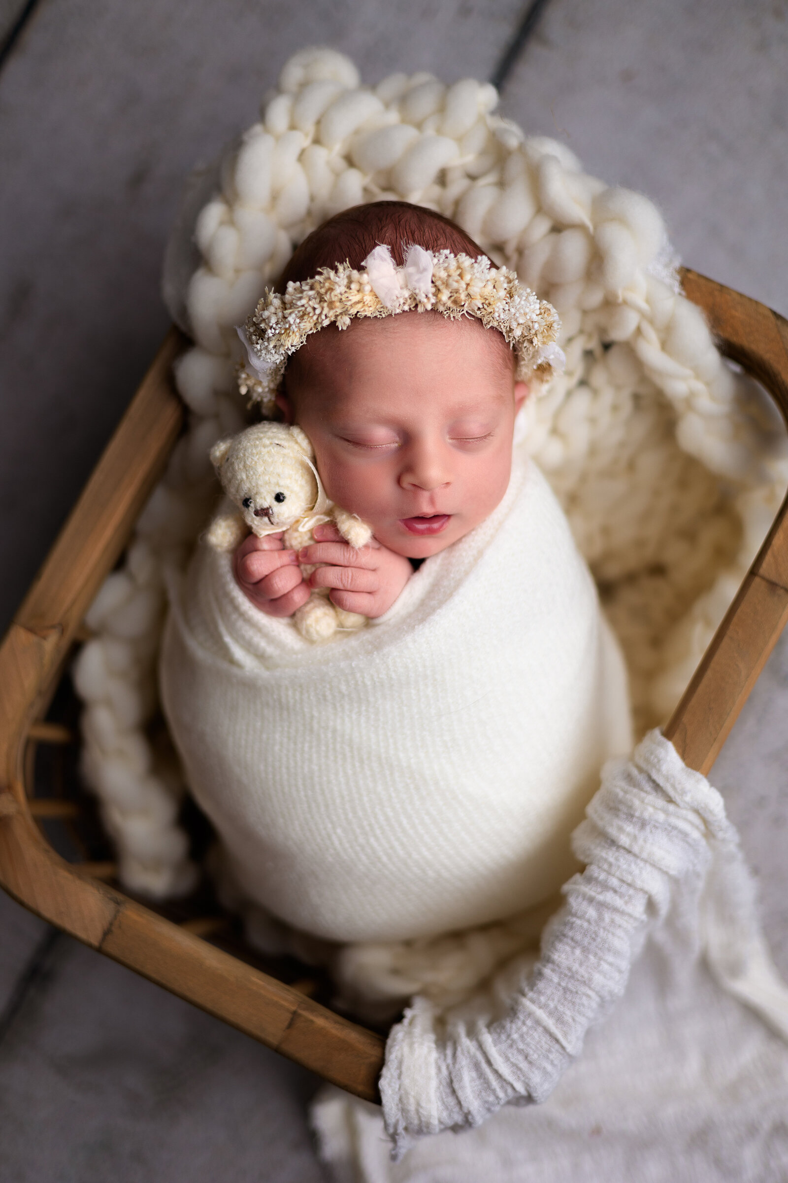 Newborn girl swaddled in a cream wrap snuggled up with a white teddy bear in a brown crate