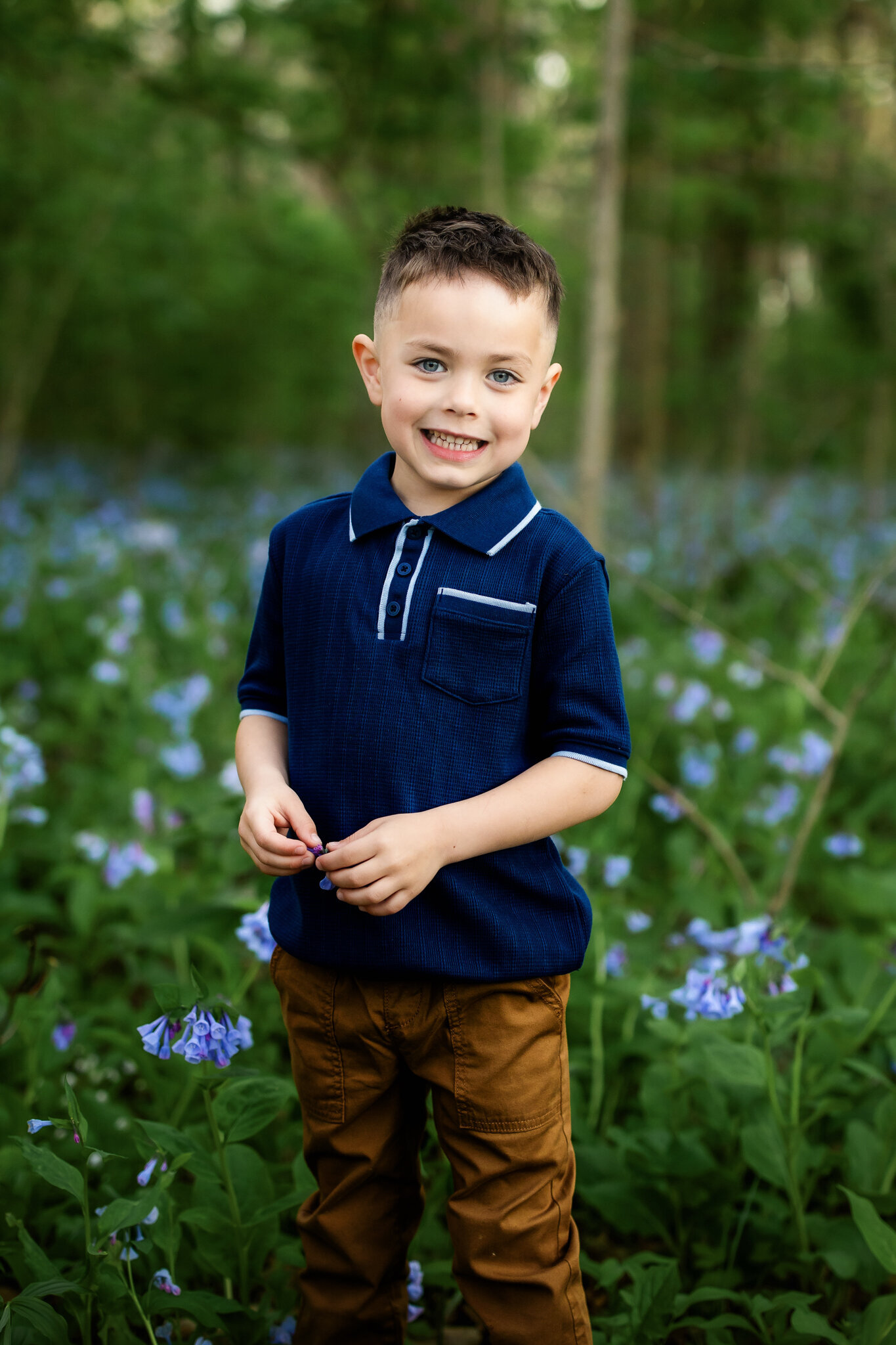 A young boy with short brown hair smiles while standing in a field of green plants and purple flowers, perfect for outdoor portraits. He wears a navy blue polo with a pocket and light brown pants. Trees and greenery blur softly in the background.