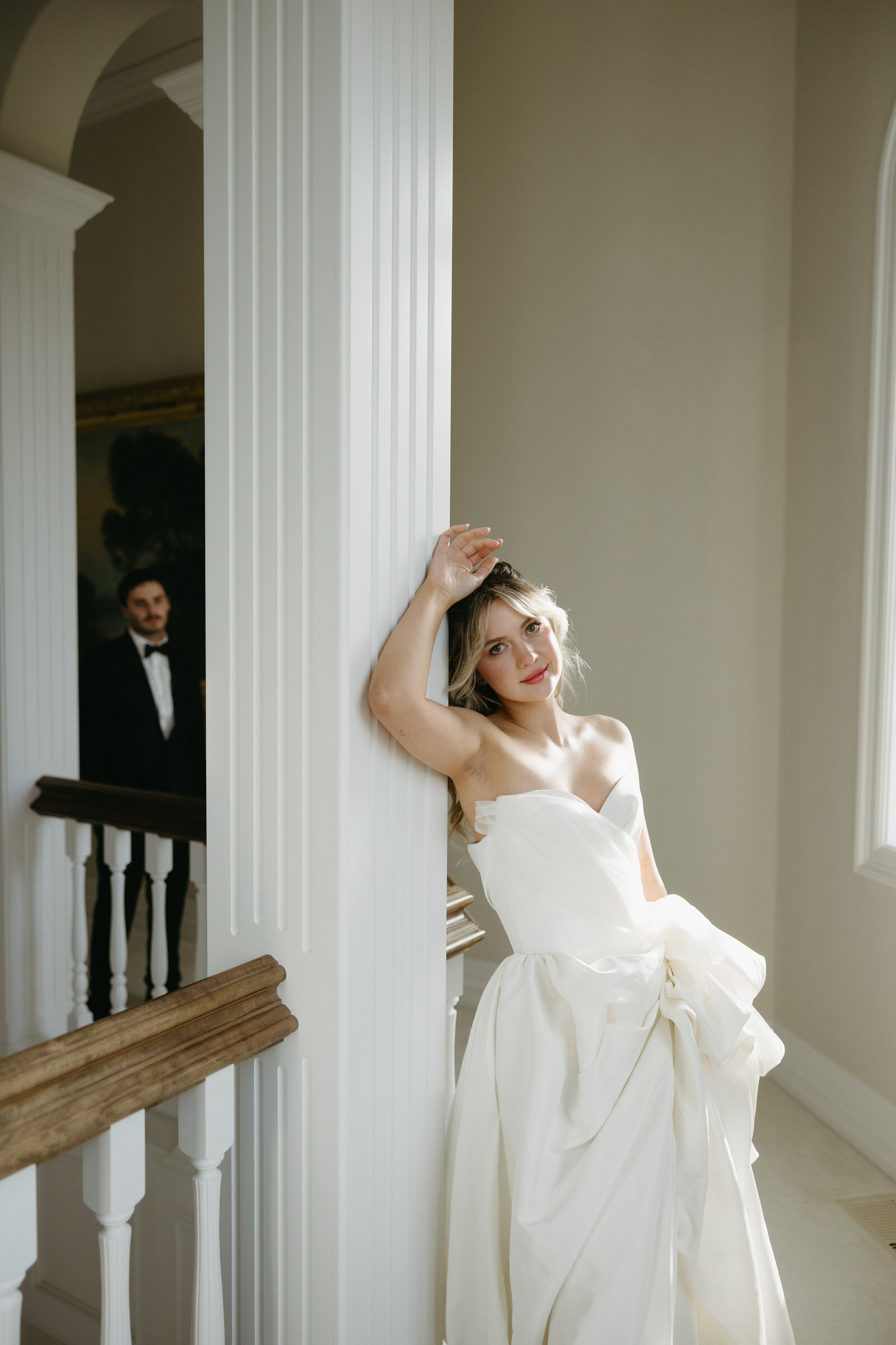 A bride is leaning on a pillar while her groom stands back to admire her. 