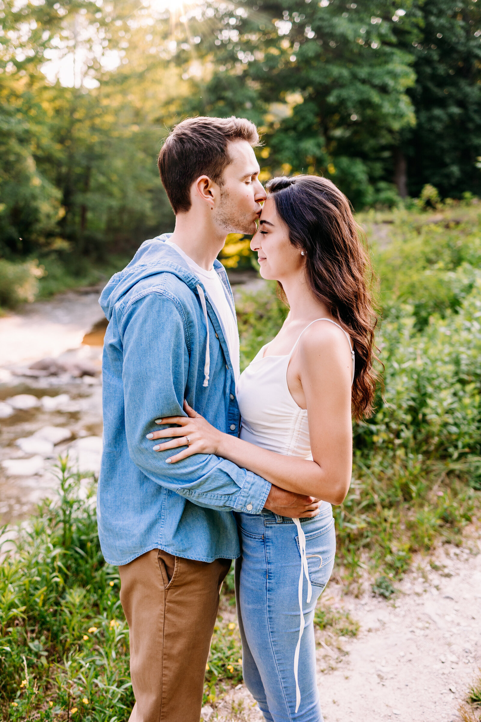 Man kisses his fiance on the forehead along  side a stream on a warm and sunny day
