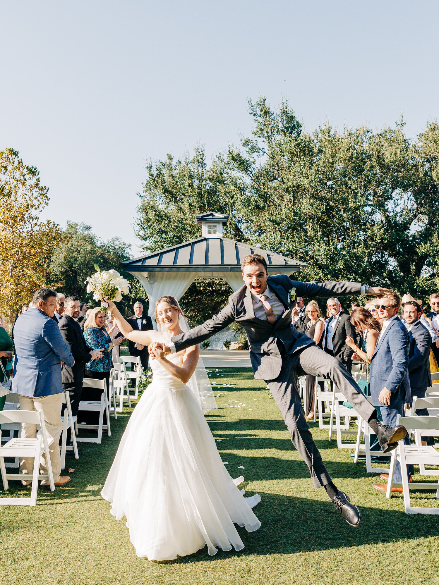 A bride and groom walk down an outdoor aisle as newlyweds; the bride smiles holding a bouquet, while the groom joyfully jumps in the air. Guests clap and celebrate under a clear, sunny sky—captured by a talented Eau Claire Wedding Photographer.