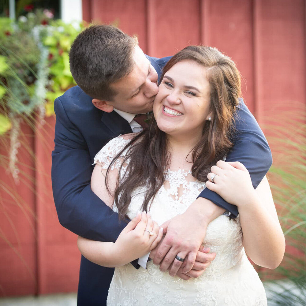 groom kisses bride's cheek in front of red building