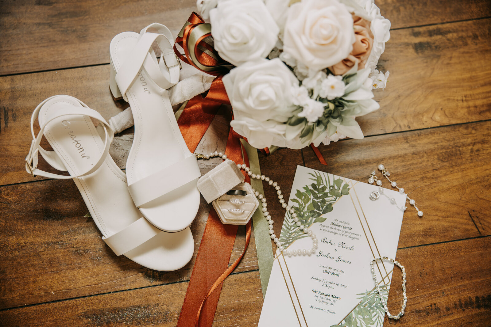 Wedding flat lay in warm fall tones of burnt orange and sage green, featuring engagement rings, ribbon, wedding invitation, bouquet, and bride's shoes.