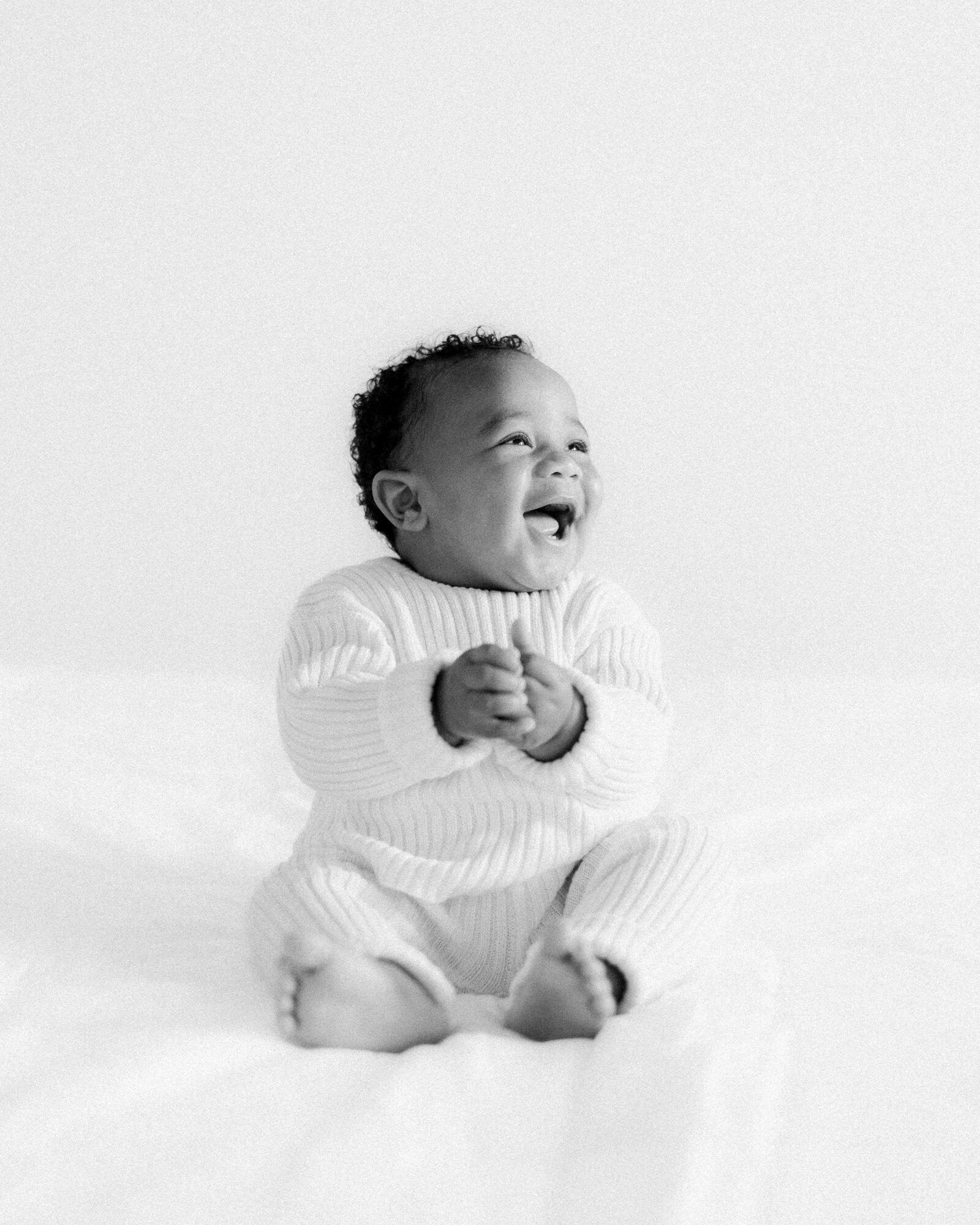 Joyful laughing one year old boy in sweater romper sitting on white bedding by NH newborn photographer Fieldstone Studio.