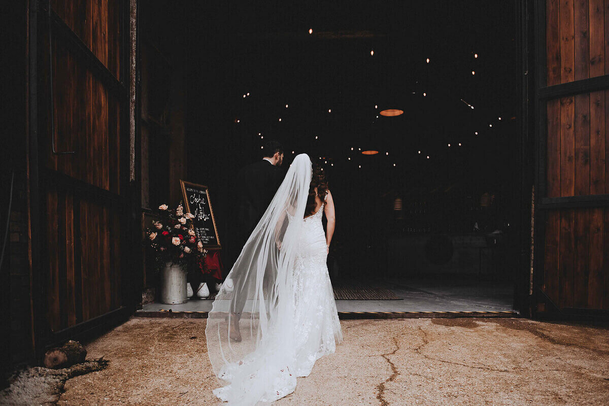 Bride and groom entering a warmly lit barn venue with a flowing veil trailing behind.