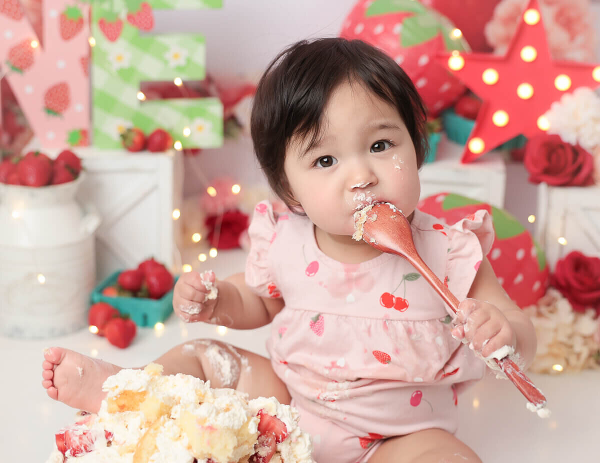 A sweet young girl licking a spoon covered in icing, surrounded by a vibrant strawberry-themed cake smash setup in a photography studio in Rochester, NY.
