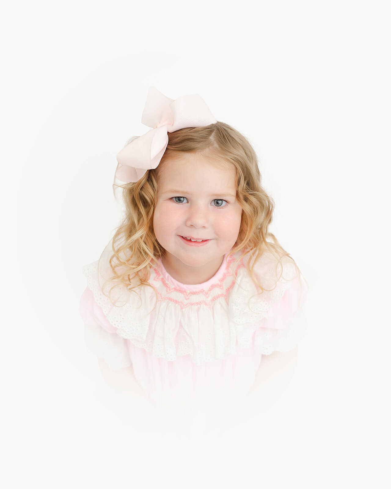A young child with curly blonde hair and a large bow in her hair, wearing a pink and white ruffled dress, gazes up at the camera. This charming portrait captures the innocence of childhood against a clean white background, showcasing the art of photography.